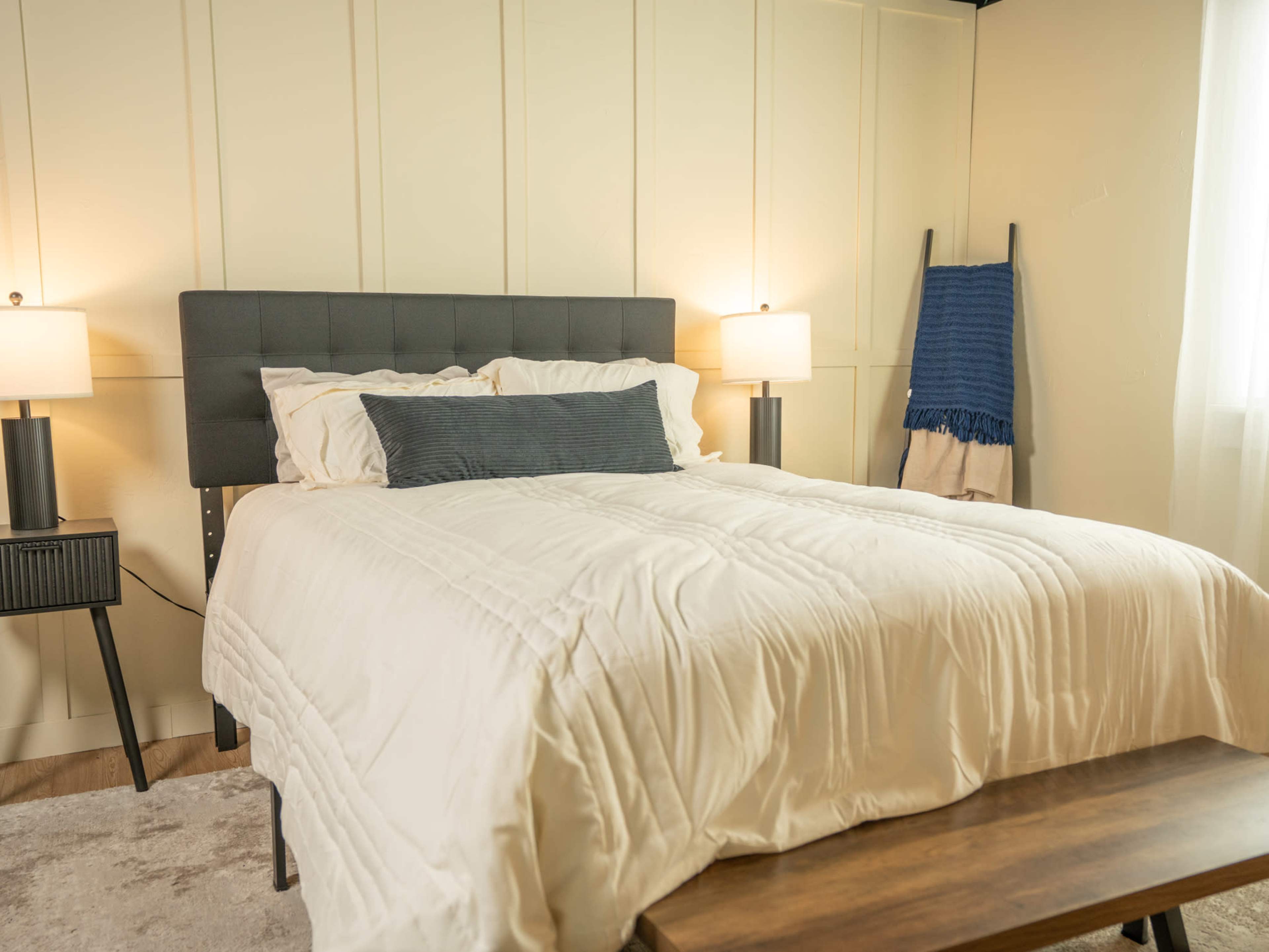 The image shows a neatly made bed with a light-colored quilt, accompanied by bedside lamps and a decorative blanket on a nearby stand in a softly lit bedroom.