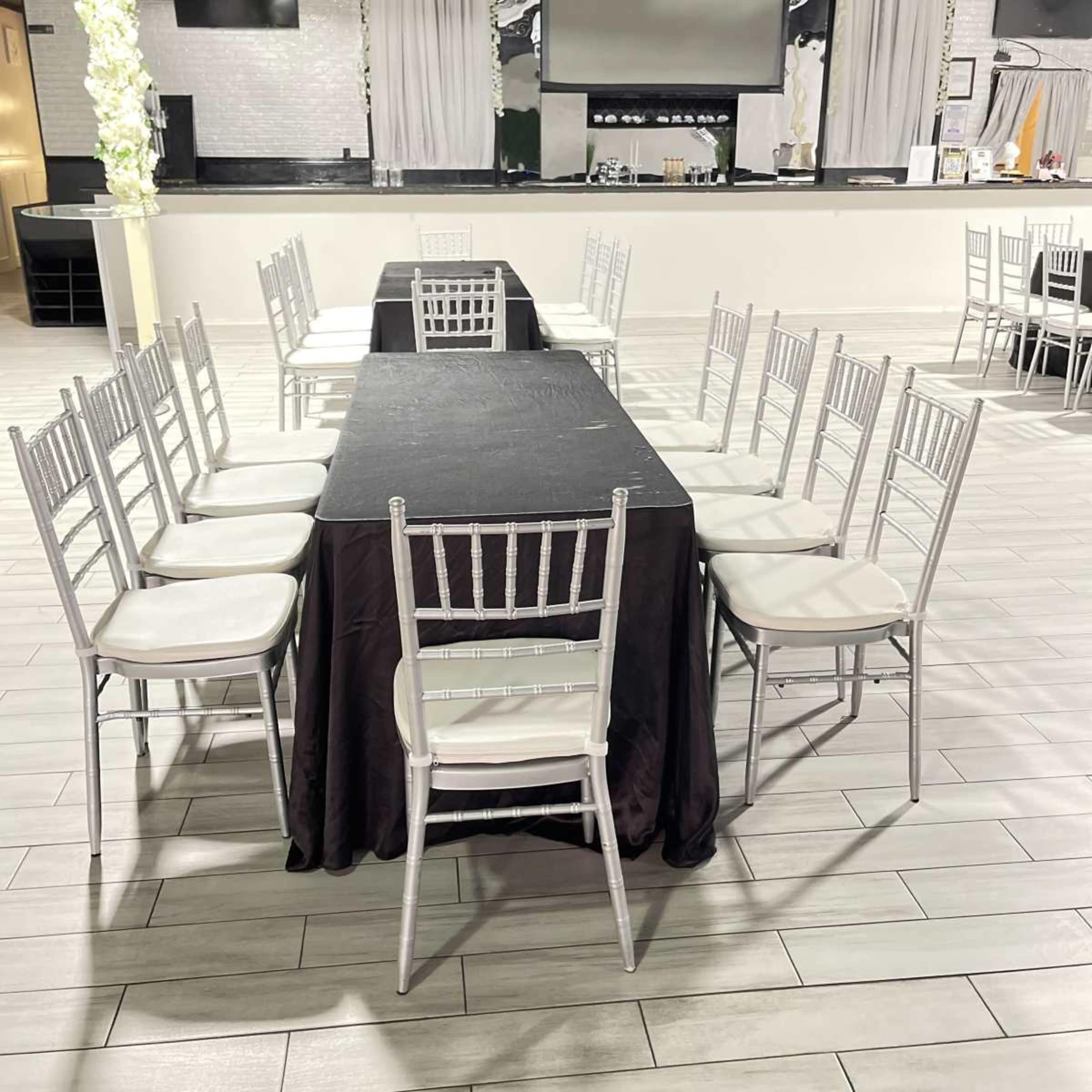 A long black table is set with silver chairs arranged around it in a spacious event venue.