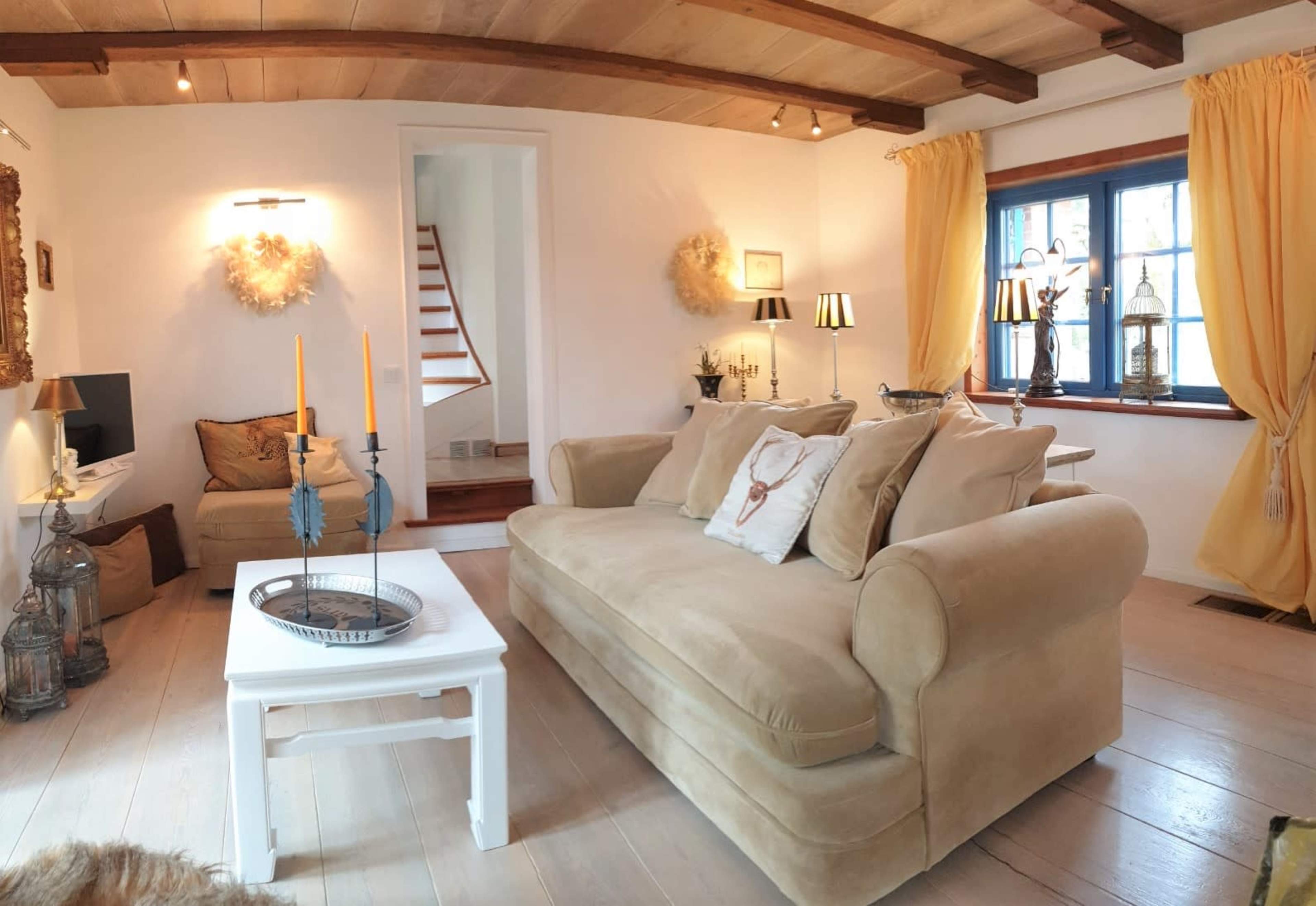 The living room features a beige sofa, a white coffee table, and warm lighting, with a staircase and large windows in the background.