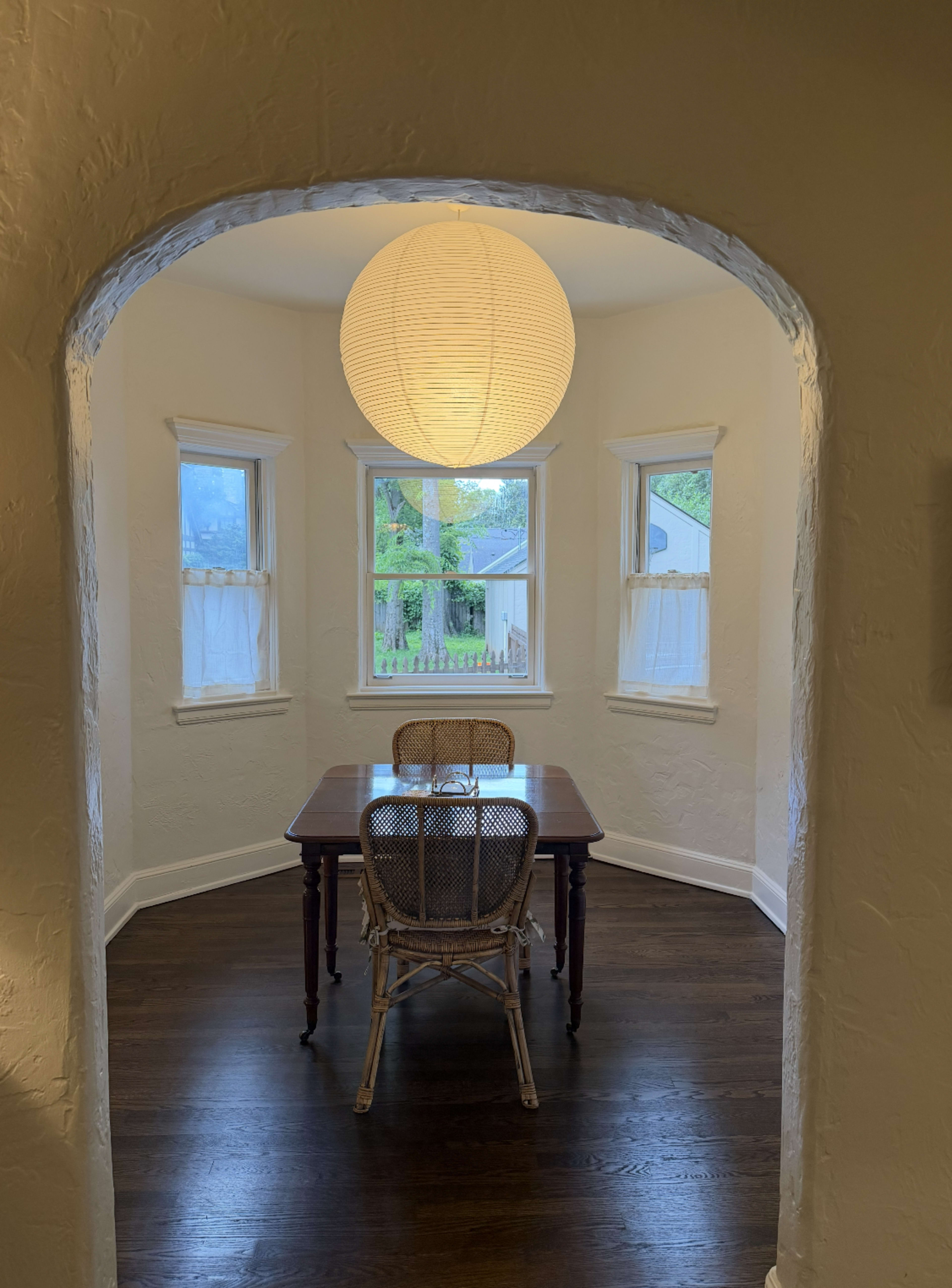 A round, white paper lantern hangs above a wooden table and chair in a bright, corner dining area with three windows.