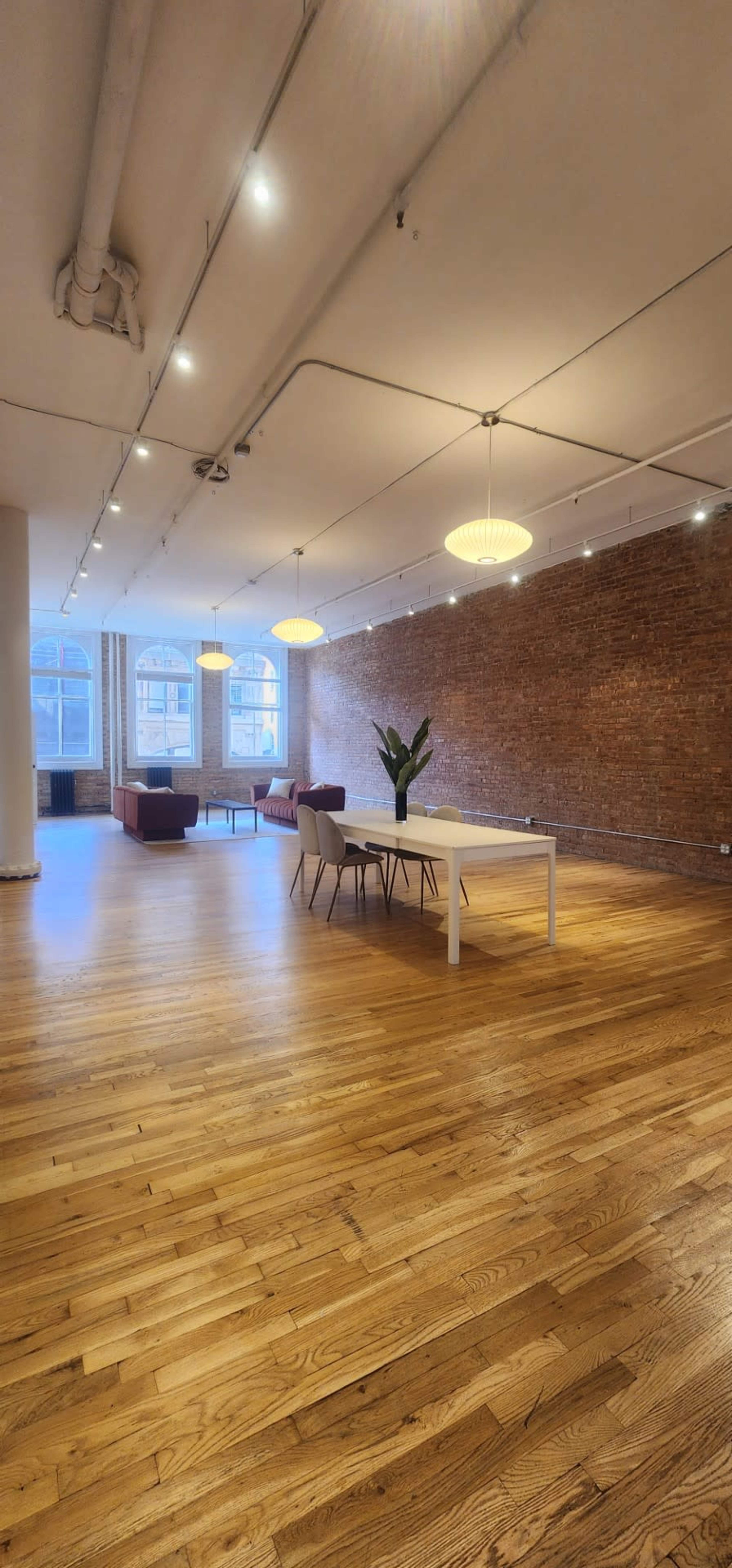 The image shows a spacious, well-lit interior featuring wooden flooring, a white dining table with chairs, a plant, and large windows with brick walls.
