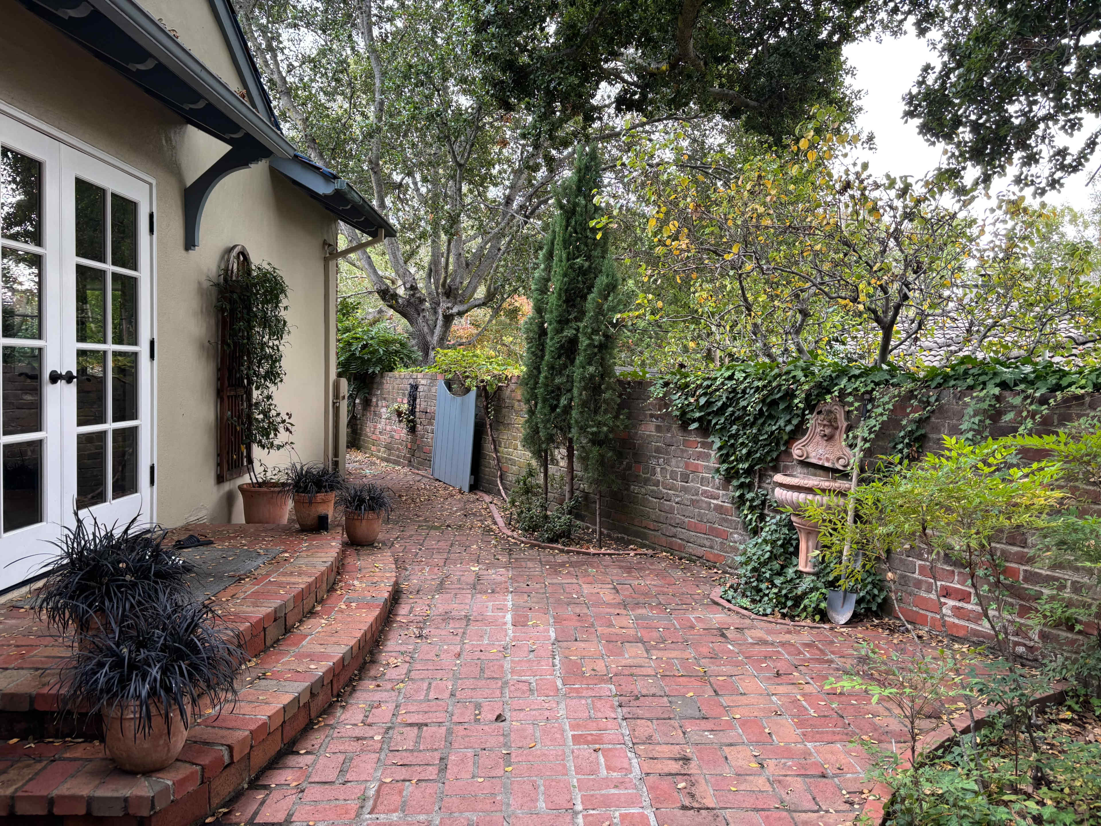 The image shows a brick pathway next to a house, flanked by potted plants and a wall covered in greenery.