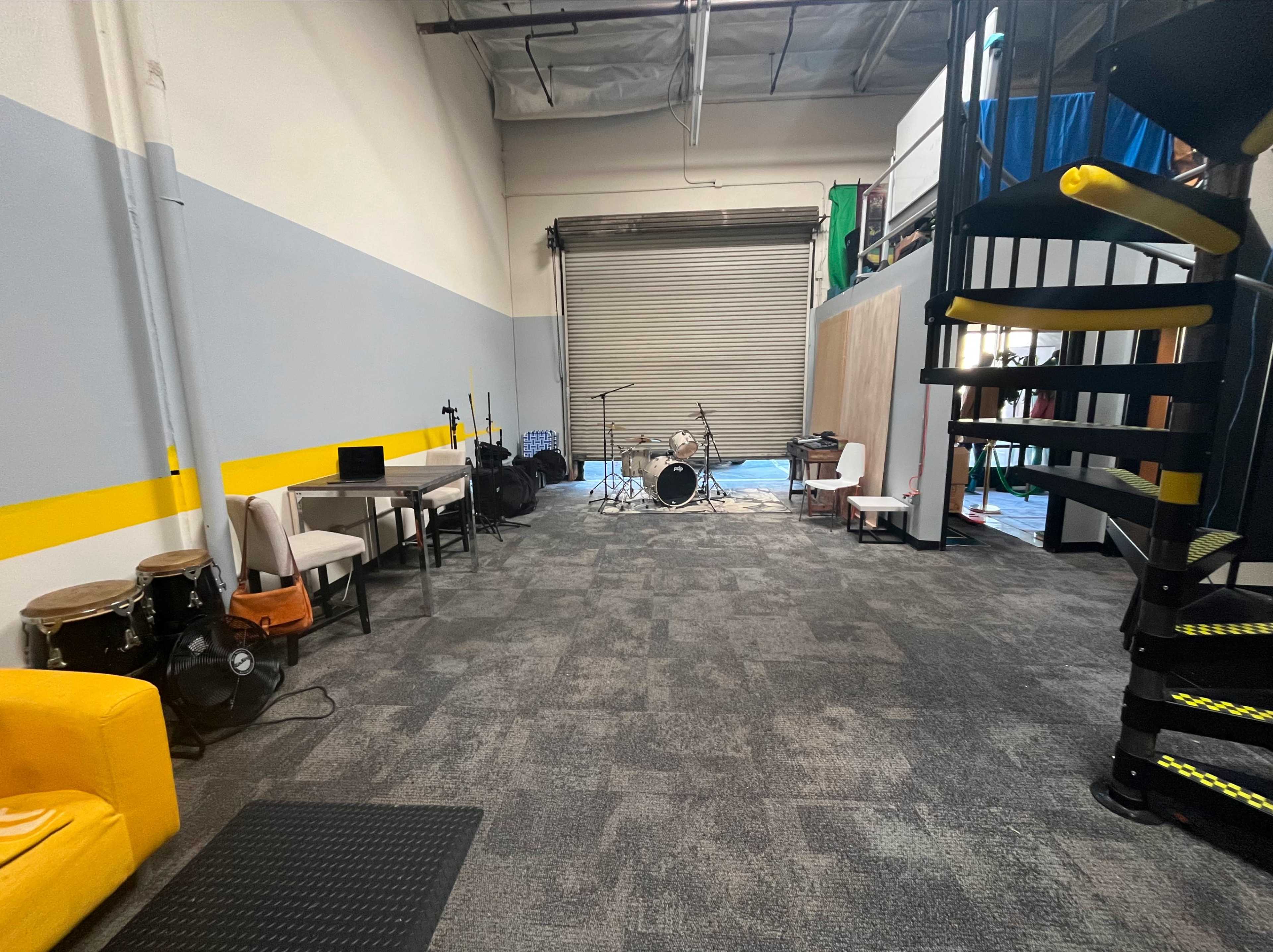 The image shows a spacious interior of a music rehearsal studio with seating, a drum set in the back, and a spiral staircase leading to an upper level.