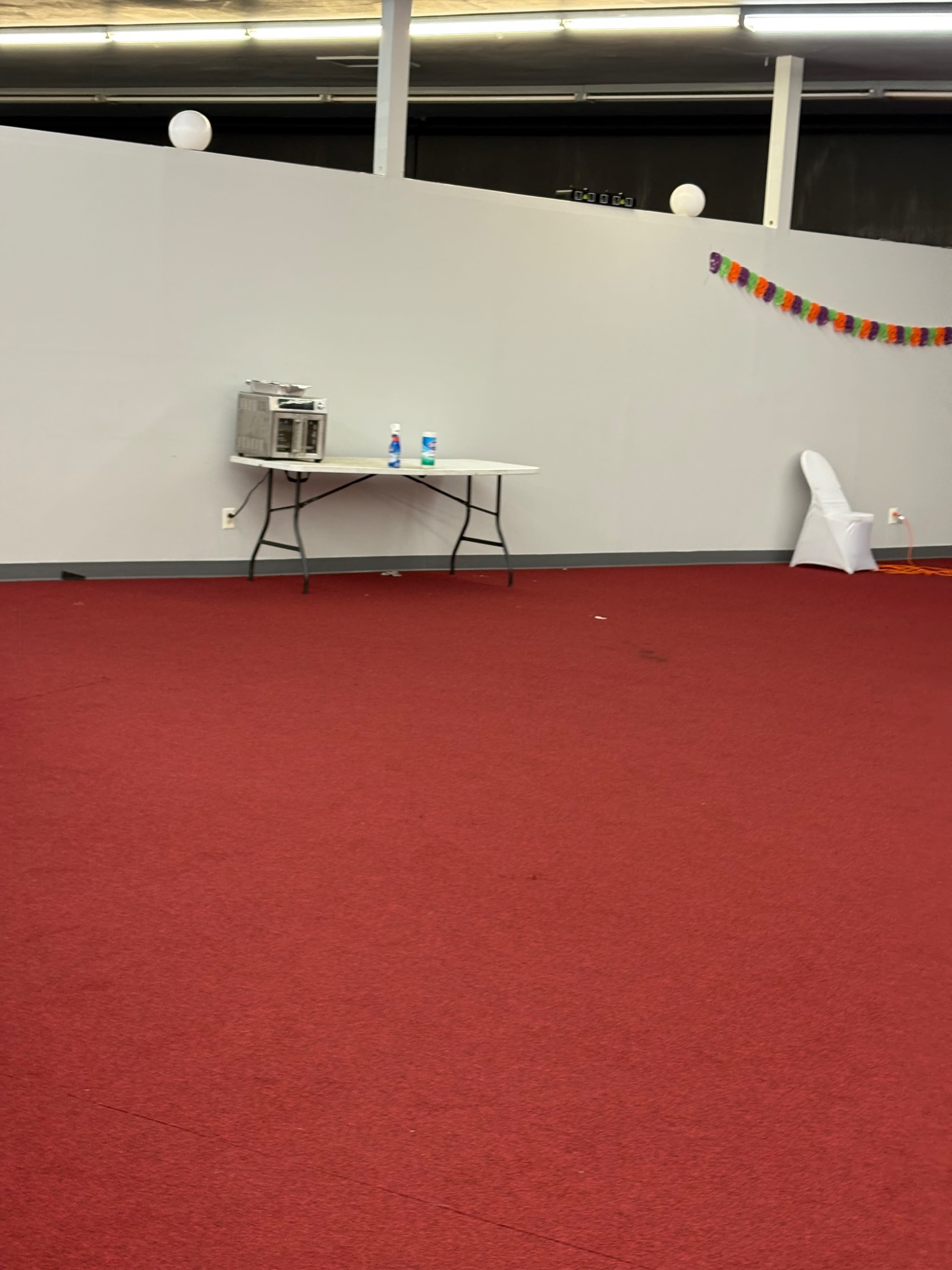 The image shows a large, empty room with red carpet, a white table holding a microwave and bottles of water, and colorful decorations along the wall.