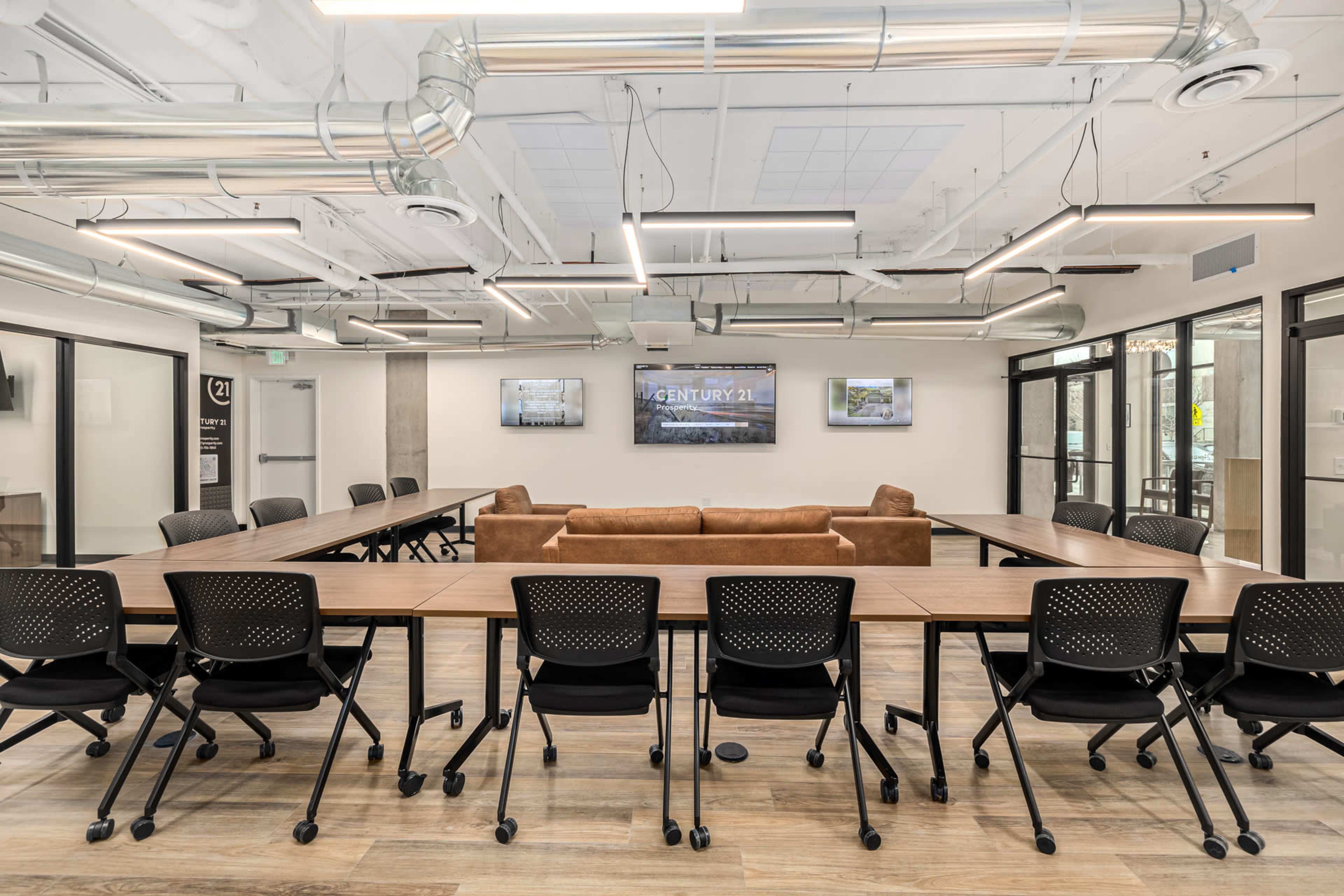 The image shows a modern conference room with long tables arranged in a U-shape, black chairs, and a brown couch, along with screens mounted on the wall.