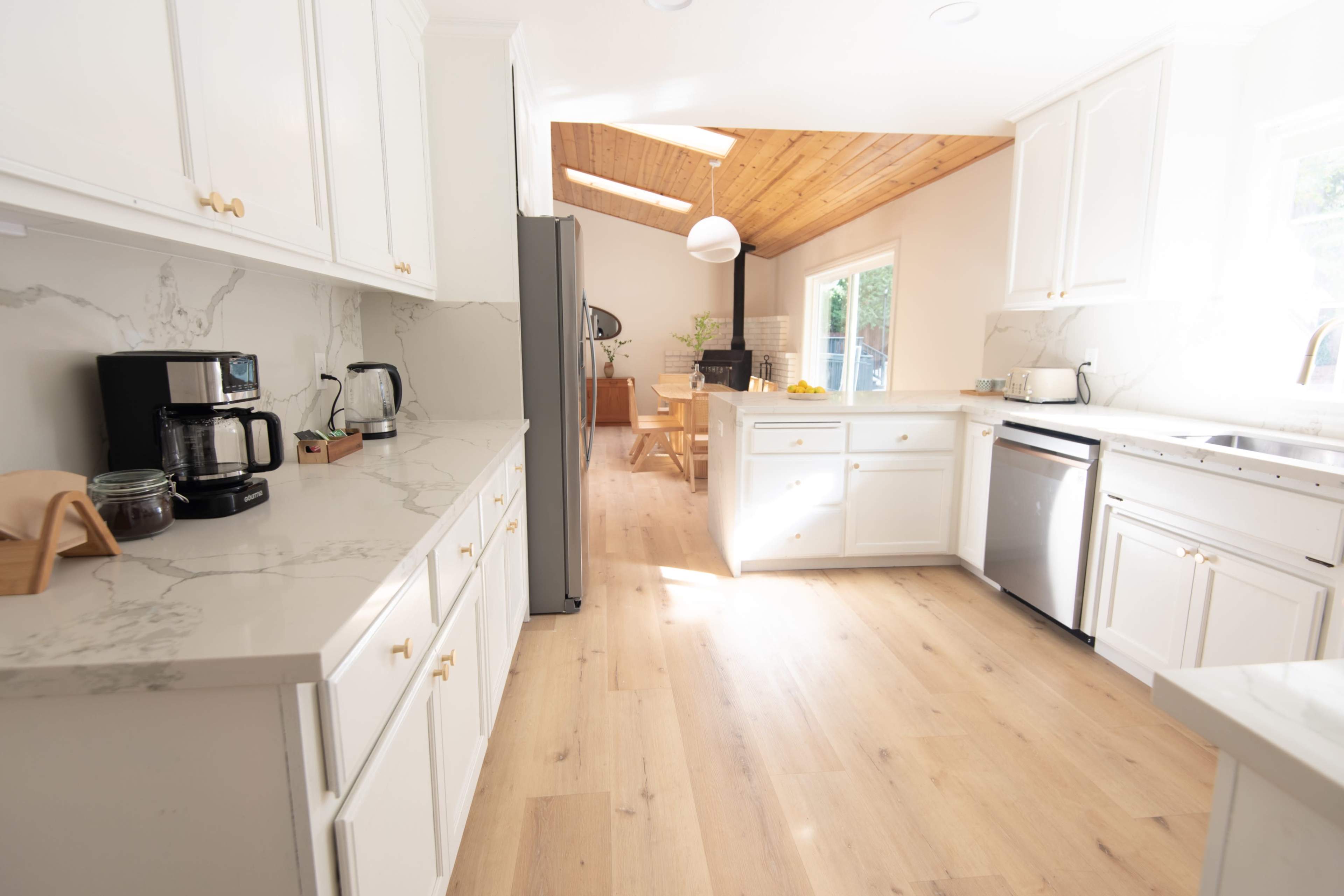 A modern kitchen with white cabinetry, a marble countertop, and light wood flooring, leading to a dining area with a table and large windows.