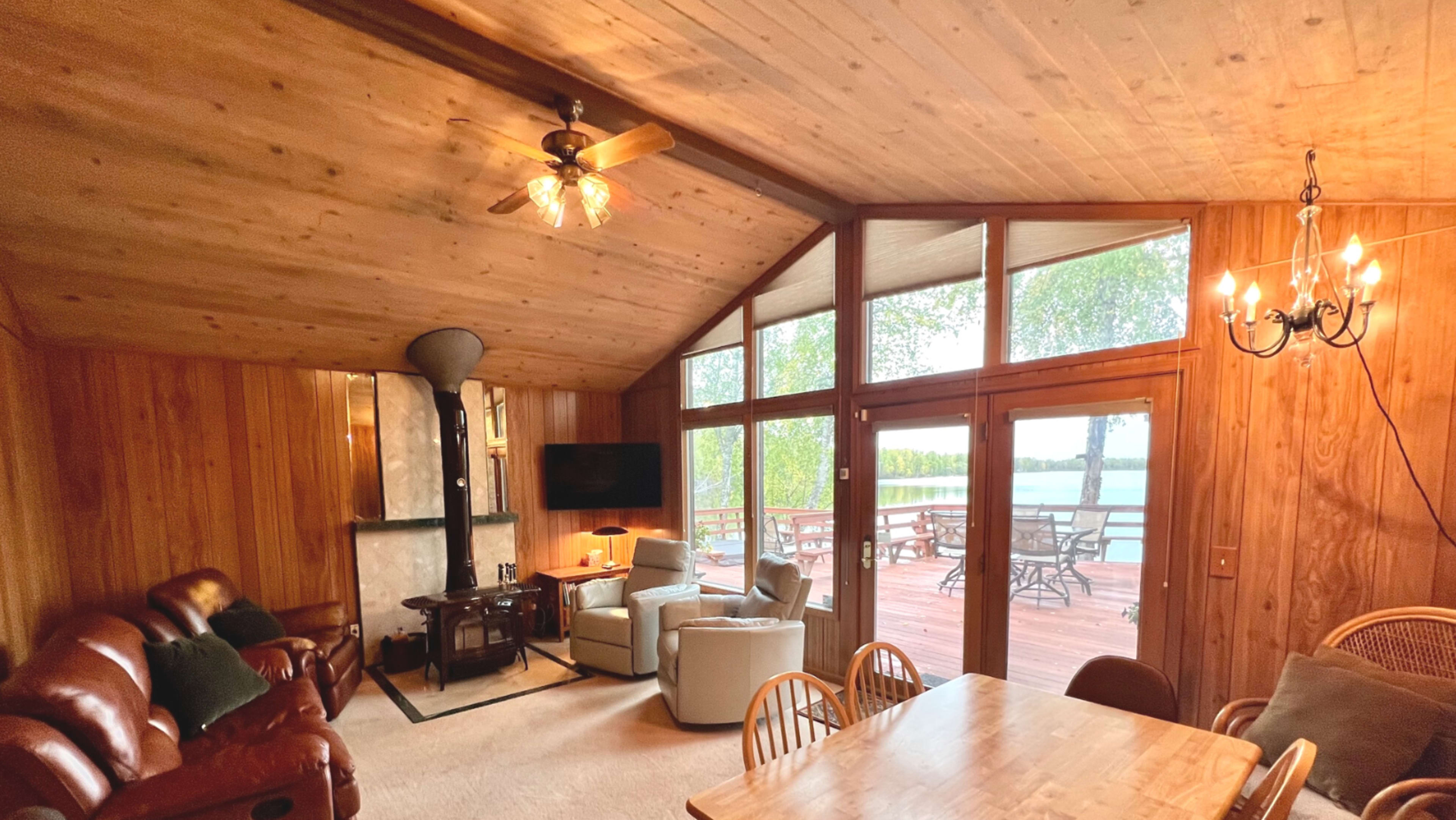 A wooden-paneled living room with a sitting area, a table, and large windows overlooking a deck and lakeside view.