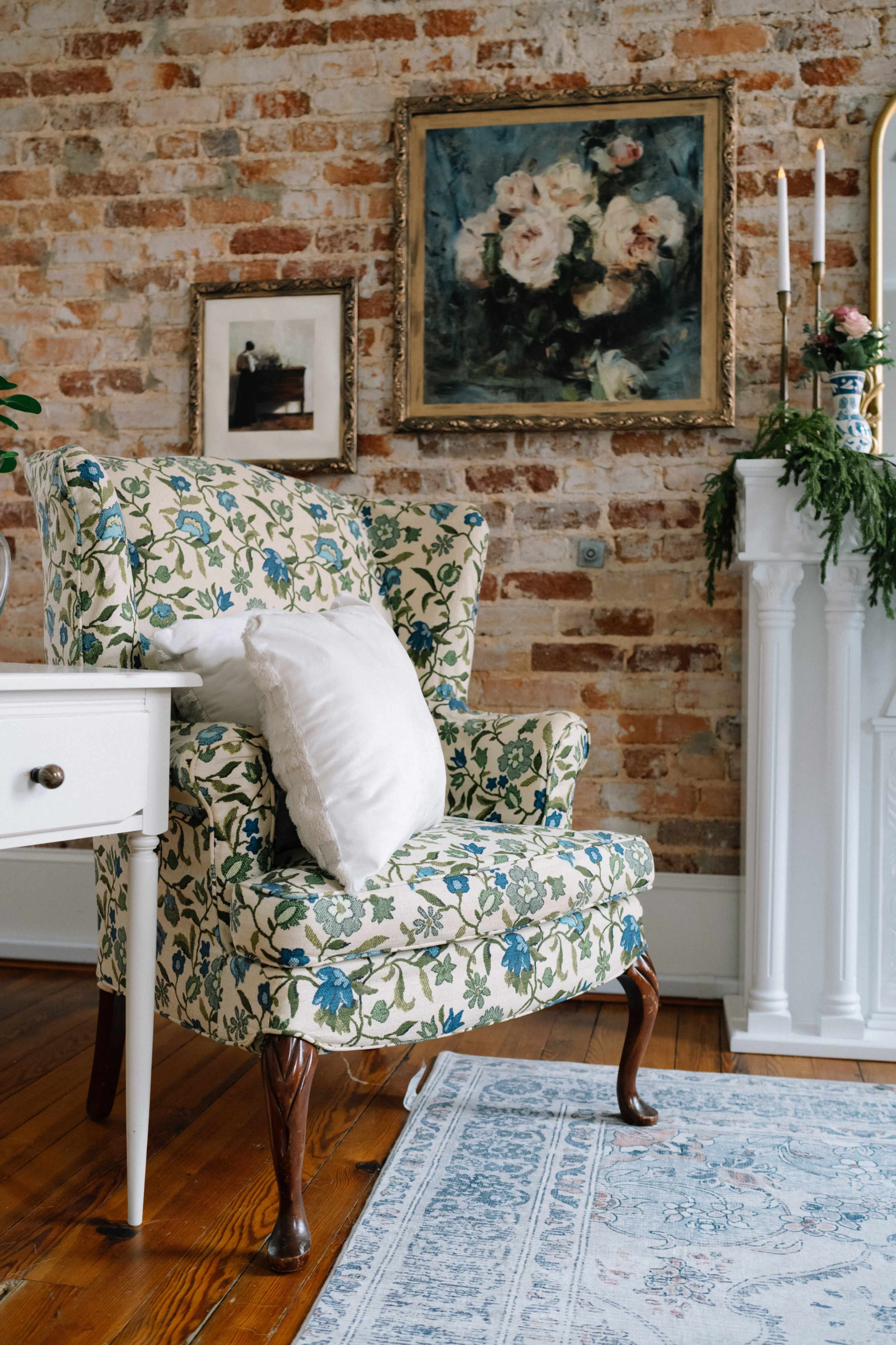 A floral-patterned armchair is positioned next to a small white table in a room with exposed brick walls and a painting of flowers above a decorative mantel.
