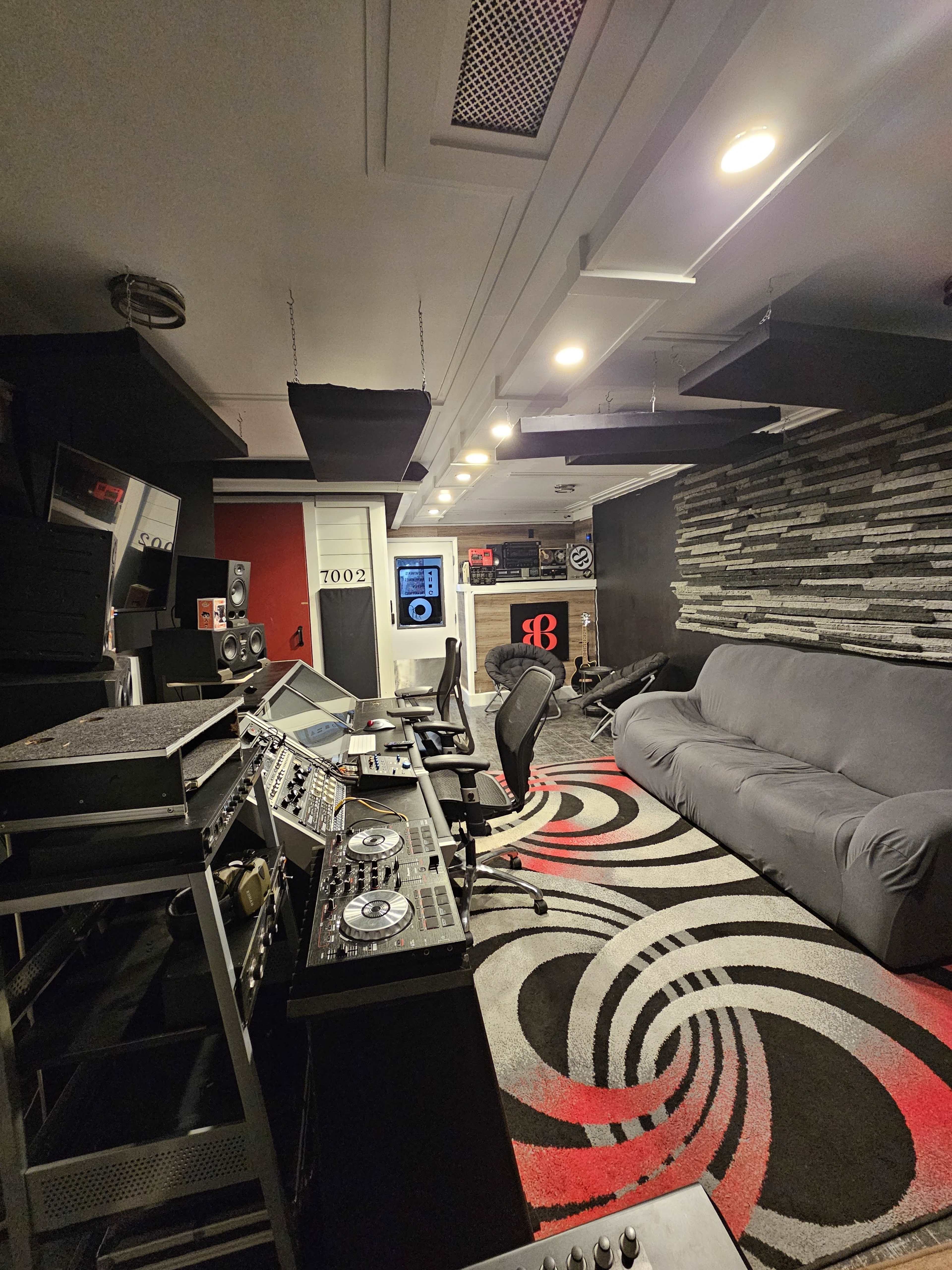 The image shows a recording studio featuring sound equipment, a large gray sofa, and patterned carpeting along with acoustic panels on the ceiling and walls.