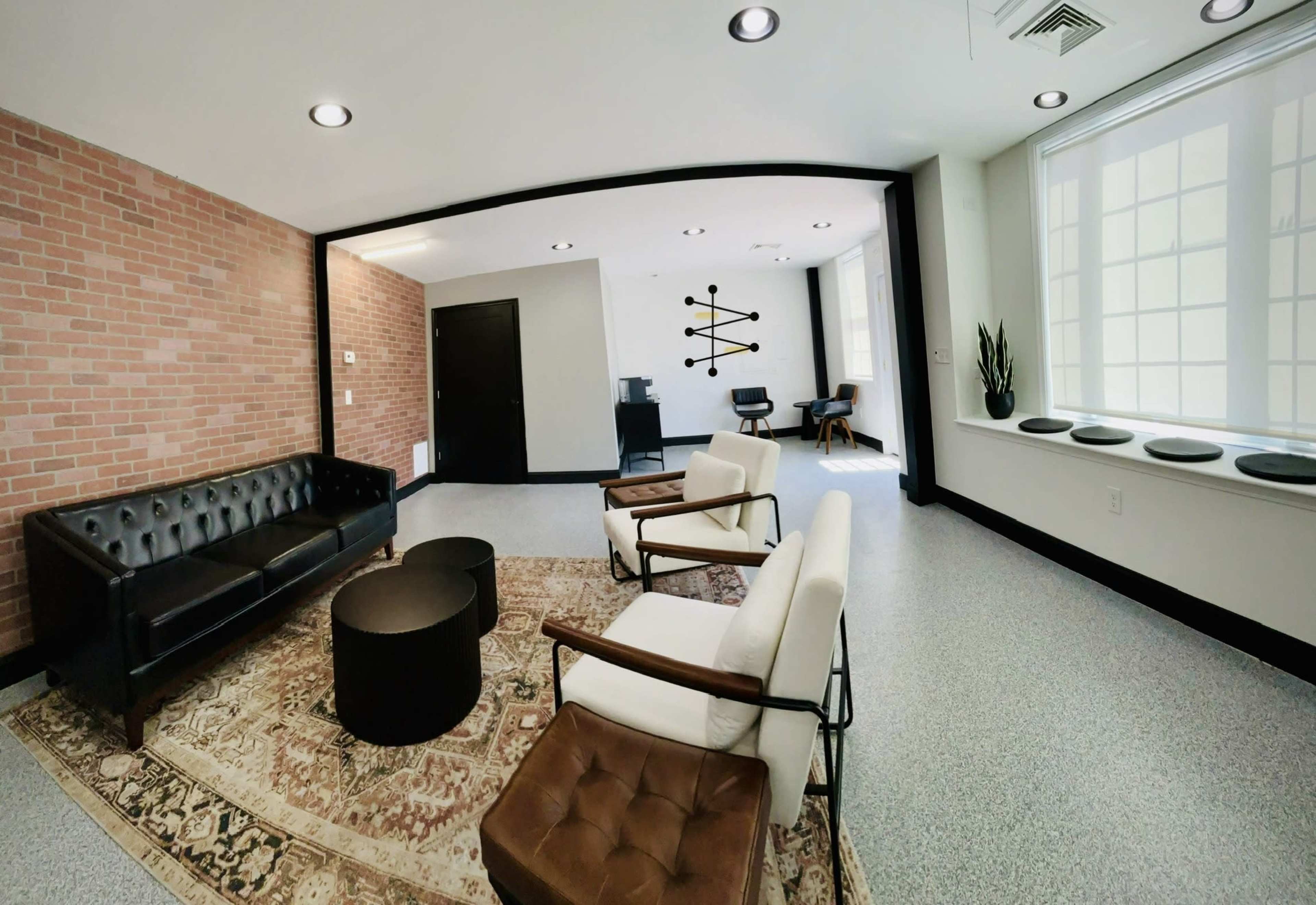 The image shows a modern lounge area featuring a black leather sofa, two white armchairs, a round black coffee table, and a patterned rug on a speckled floor.