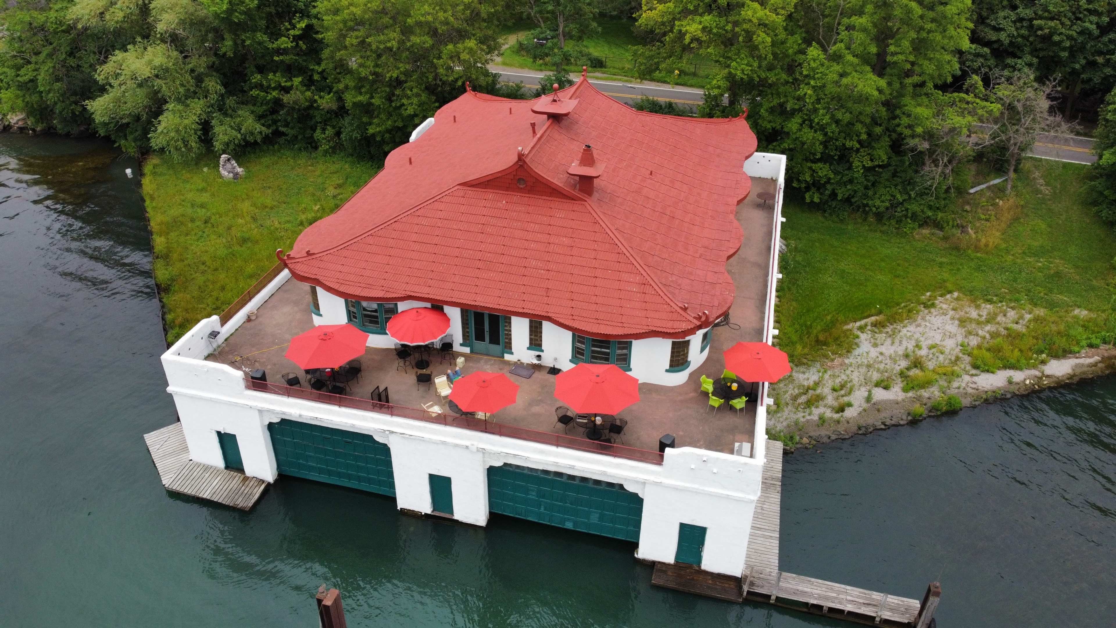 An elevated view shows a white building with a distinctive red roof, surrounded by water and featuring a patio with red umbrellas.