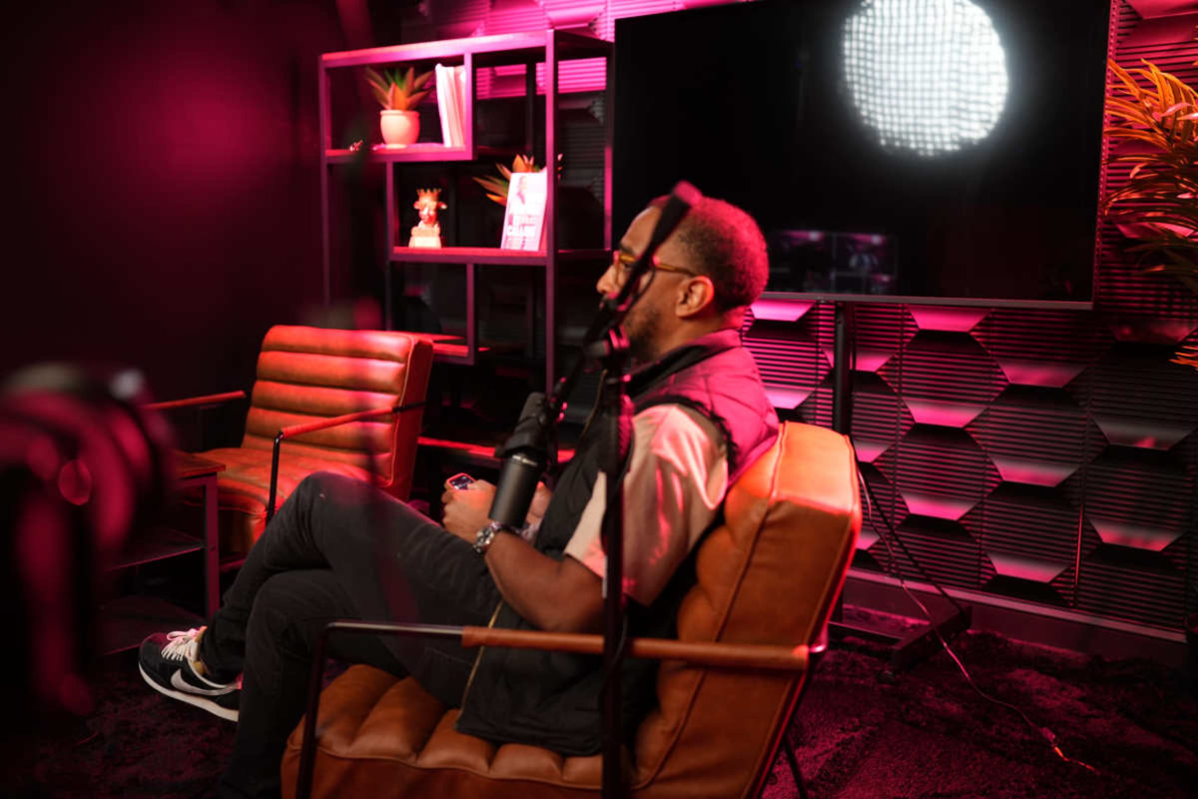 A person sits in a chair playing a video game in a modern, dimly lit studio with pink lighting.