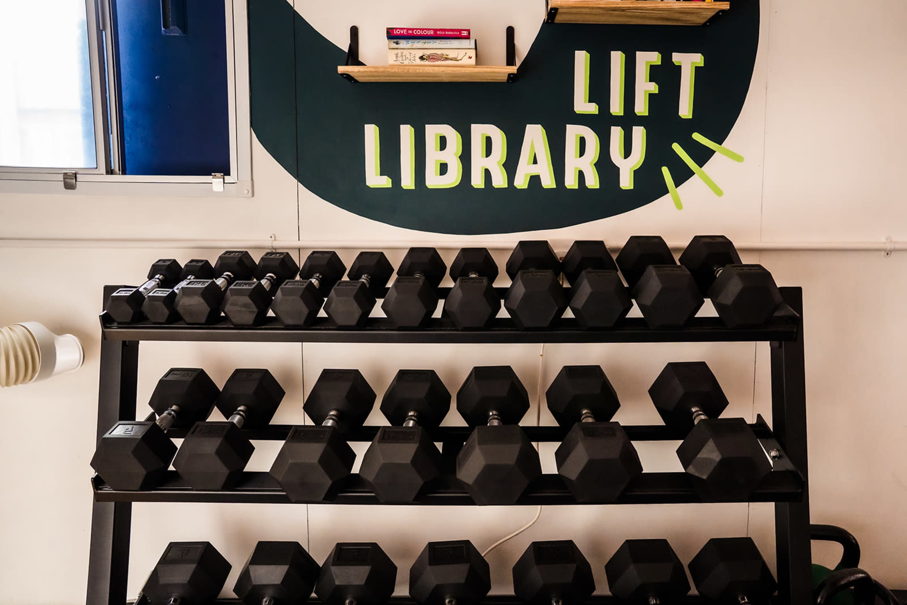 The image shows a rack of black dumbbells beneath a wall sign that reads "LIFT LIBRARY."