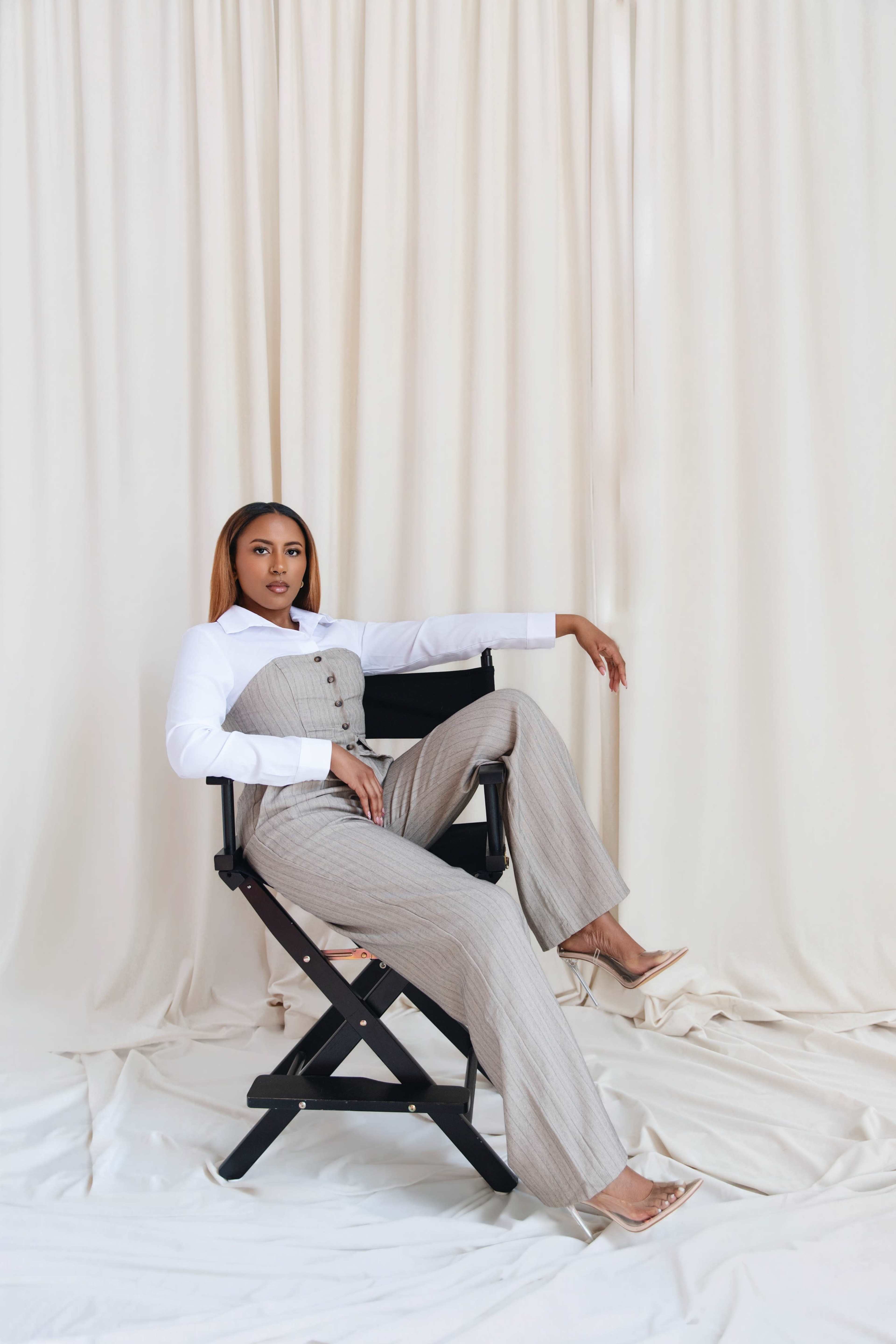 A woman in a white button-up shirt and gray striped pants sits elegantly on a director's chair in front of a plain backdrop.