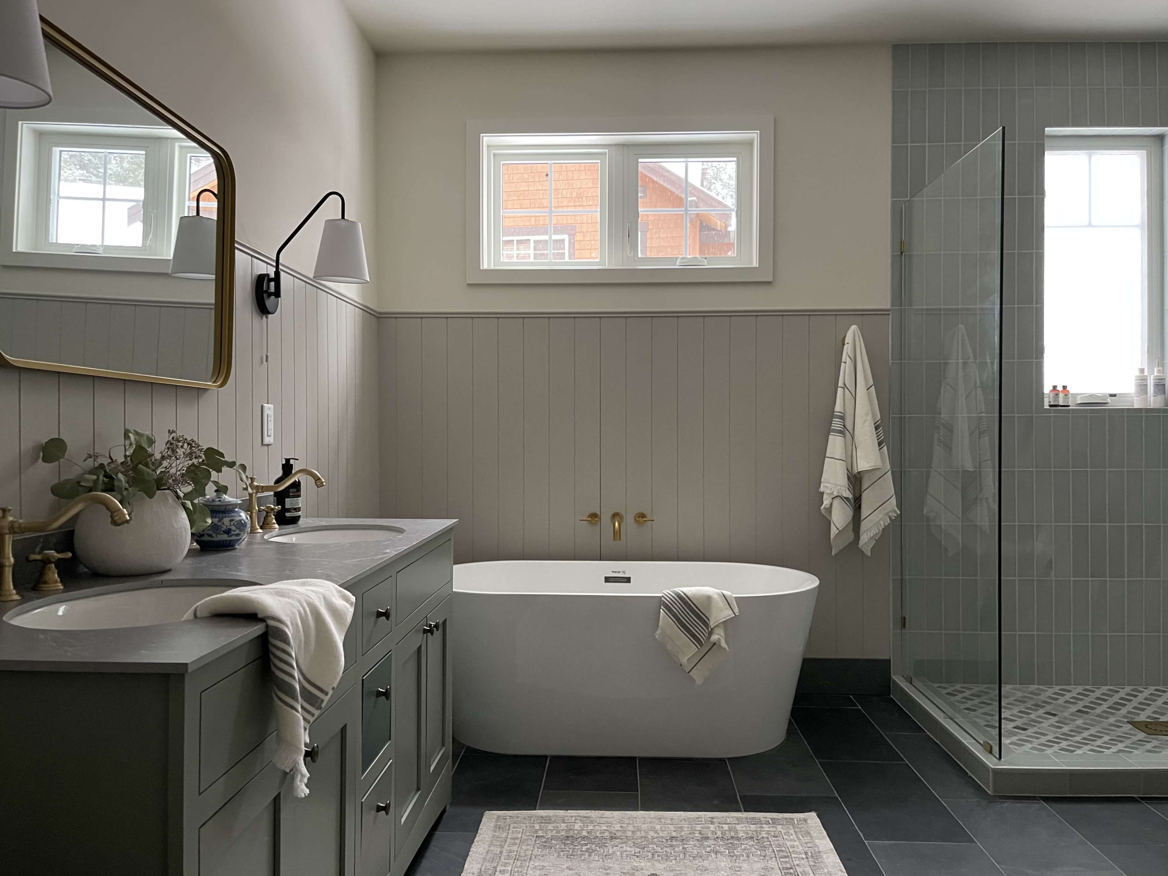 A modern bathroom features a freestanding bathtub, a glass shower stall, a double sink vanity with green cabinets, and large windows allowing natural light.