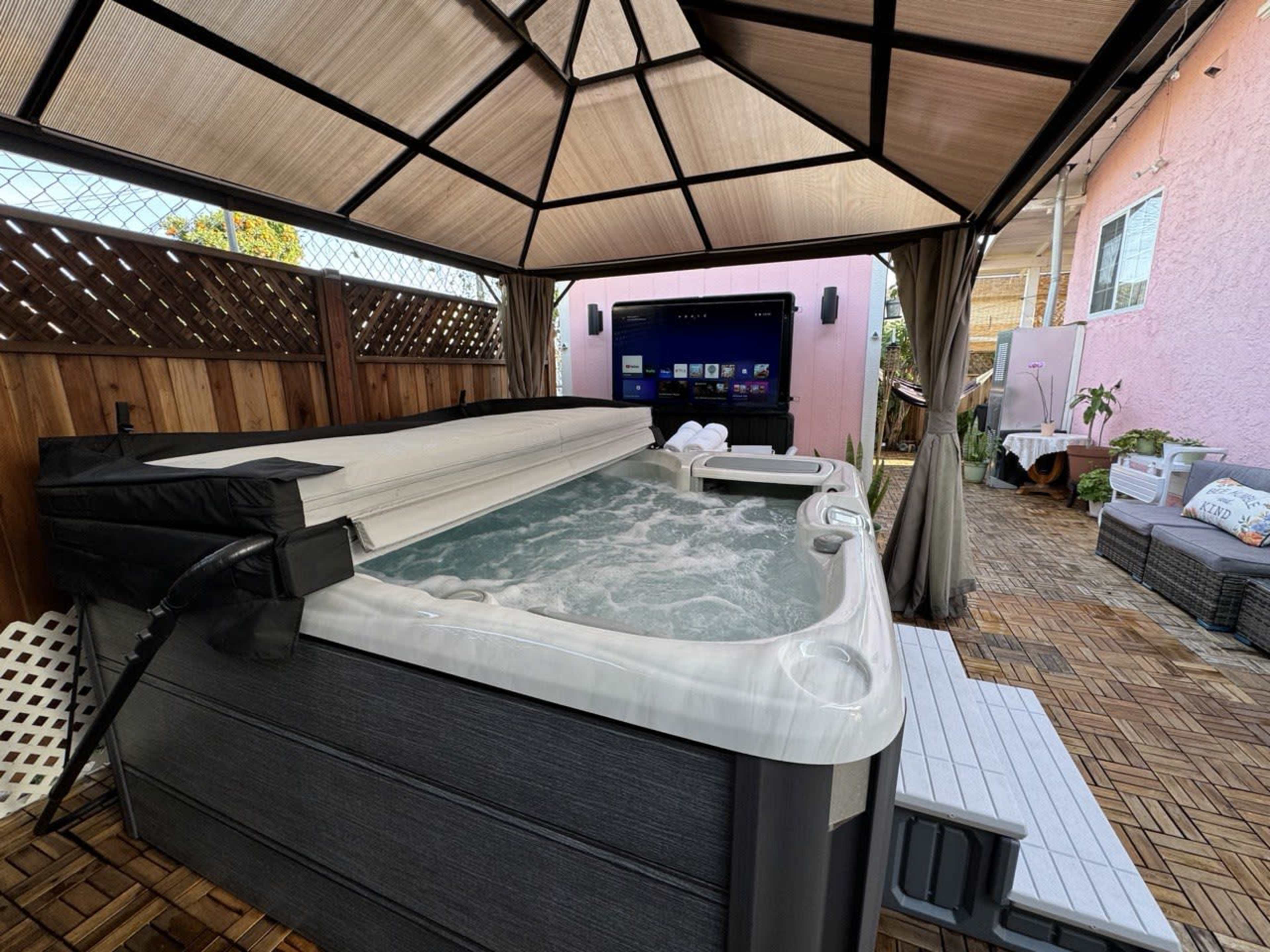 A covered outdoor area features a hot tub, a television, and seating arrangements on wooden decking.