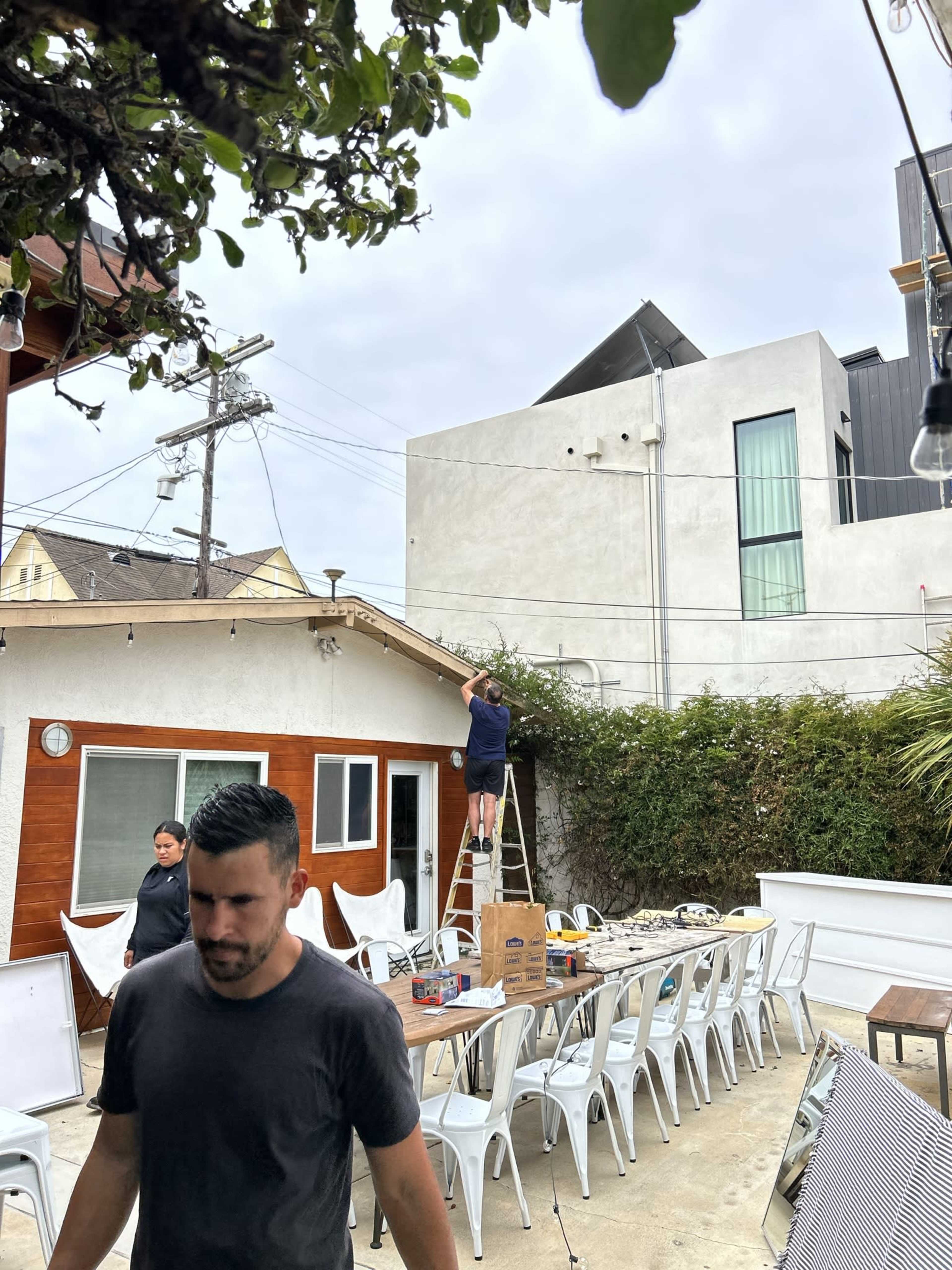 A man stands on a ladder near a house, while another man walks past in an outdoor setting with tables and chairs.