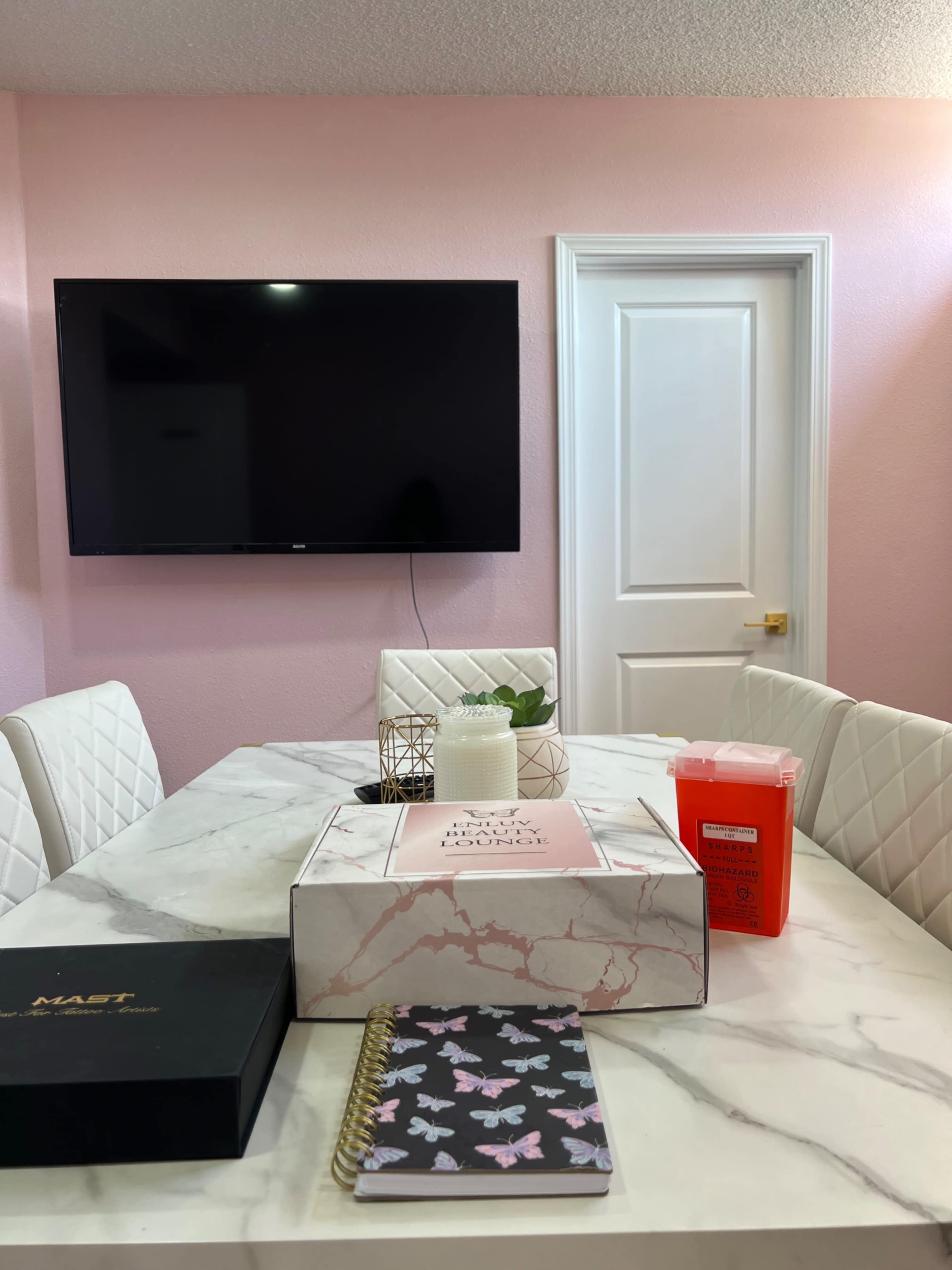 A dining table with a marble top is set in a pink-walled room, featuring a large TV on the wall, a candle, a planner, and a decorative box.