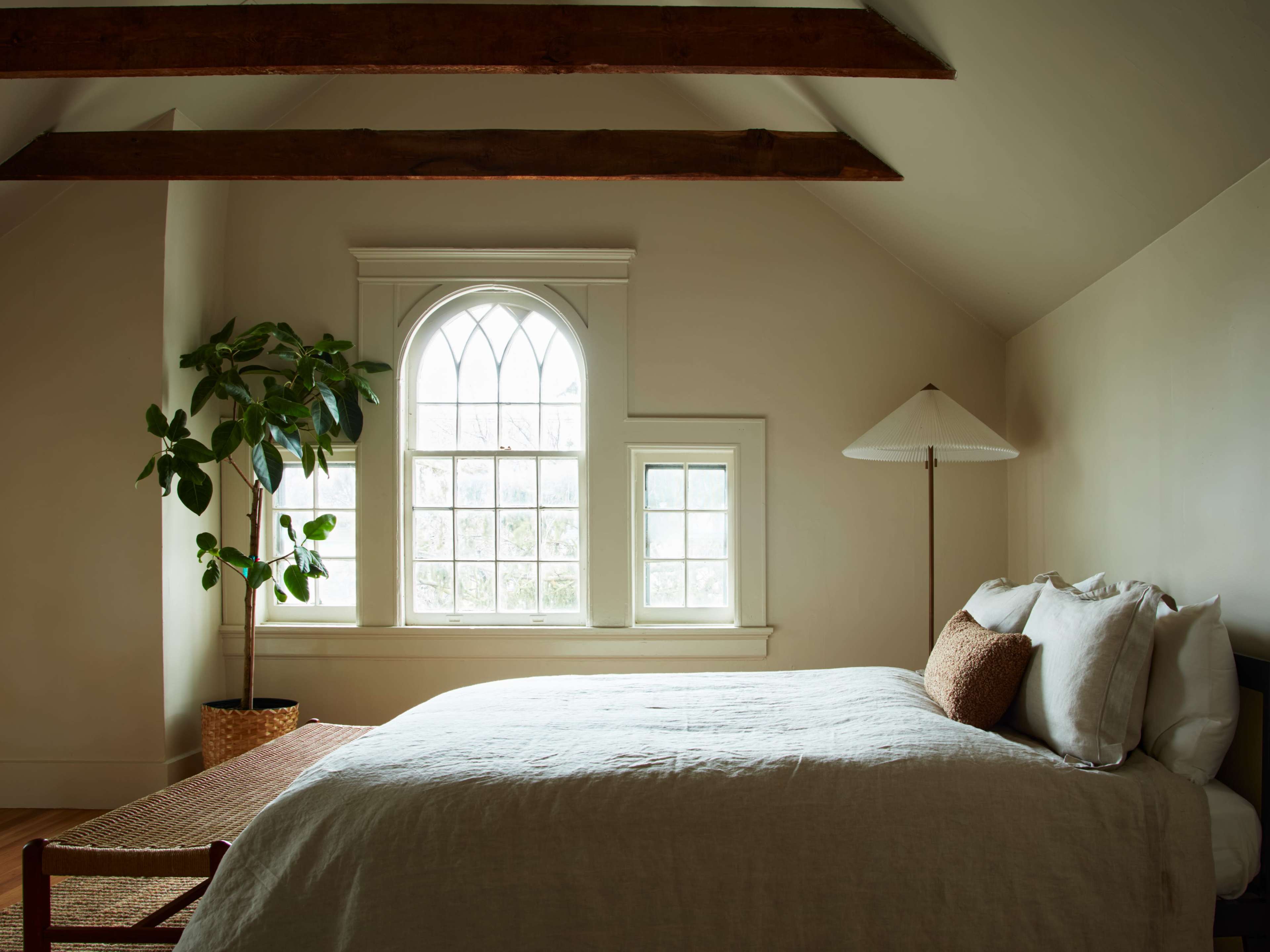 A well-lit bedroom features a large window with an arch design, a bed with neutral bedding, a potted plant, and a floor lamp.