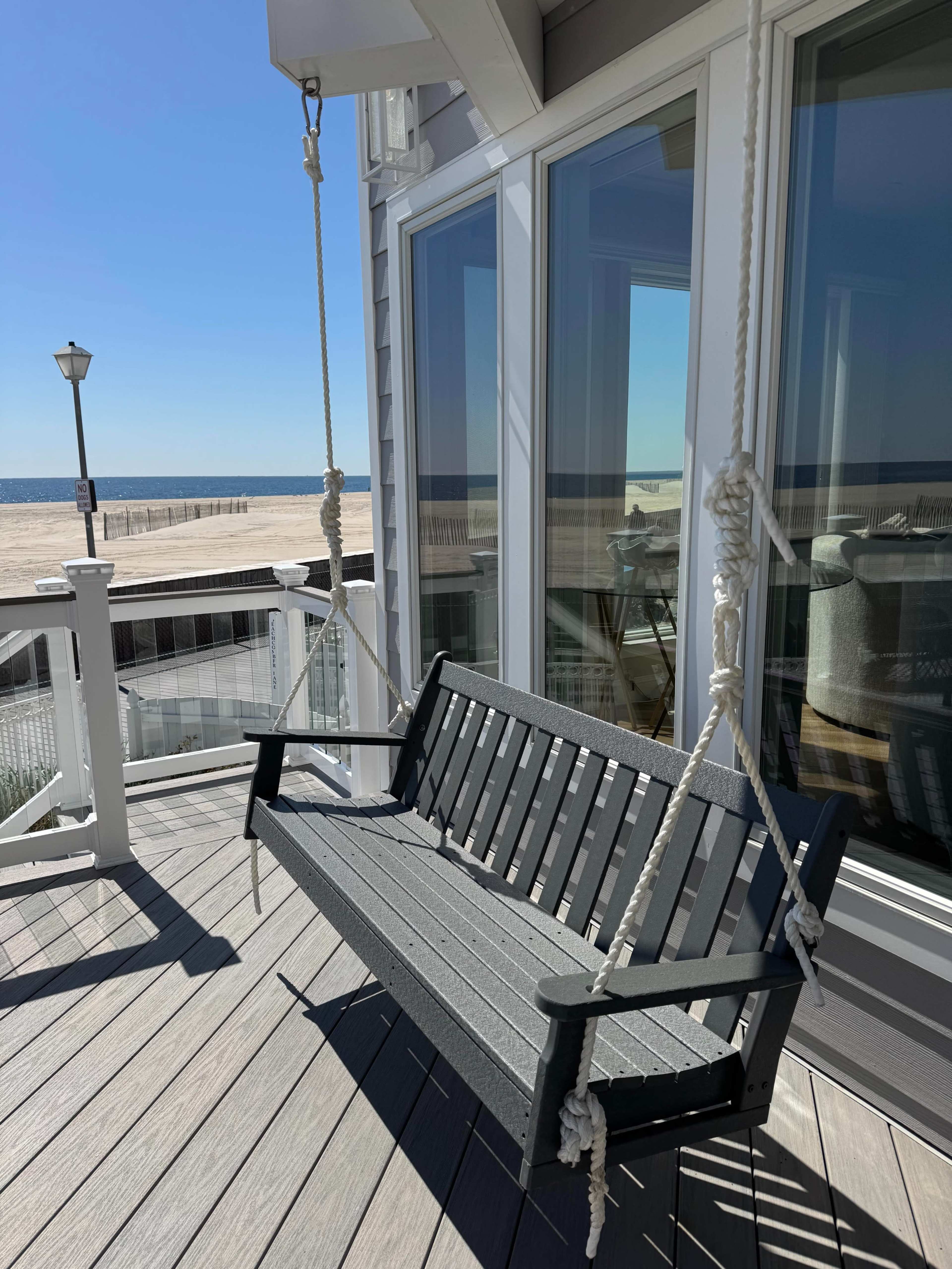 A swing bench is suspended on ropes outside a beachside house, with a view of the sandy beach and ocean in the background.