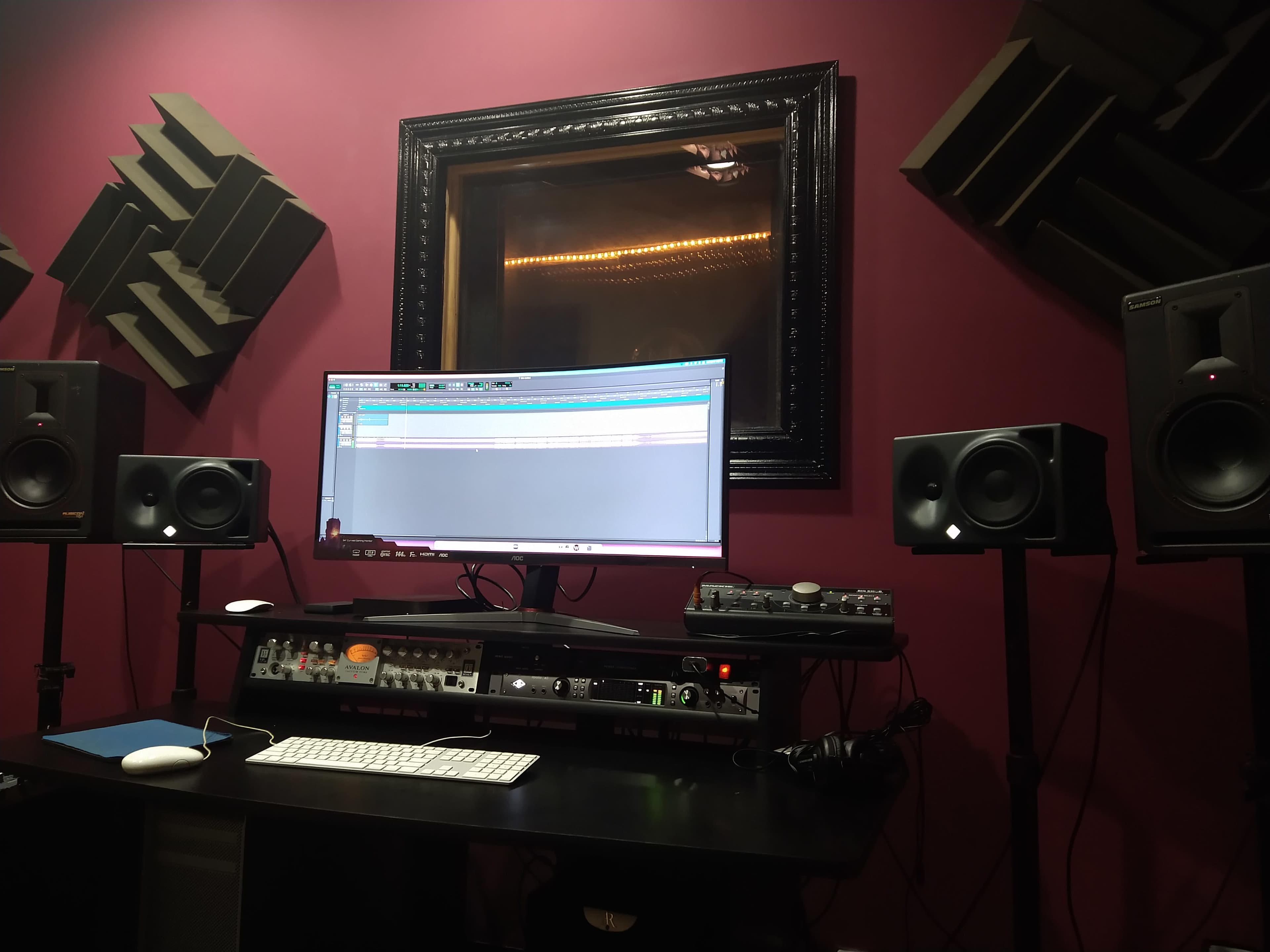 A music production studio featuring a monitor, audio speakers, and studio equipment arranged on a desk against a maroon wall.