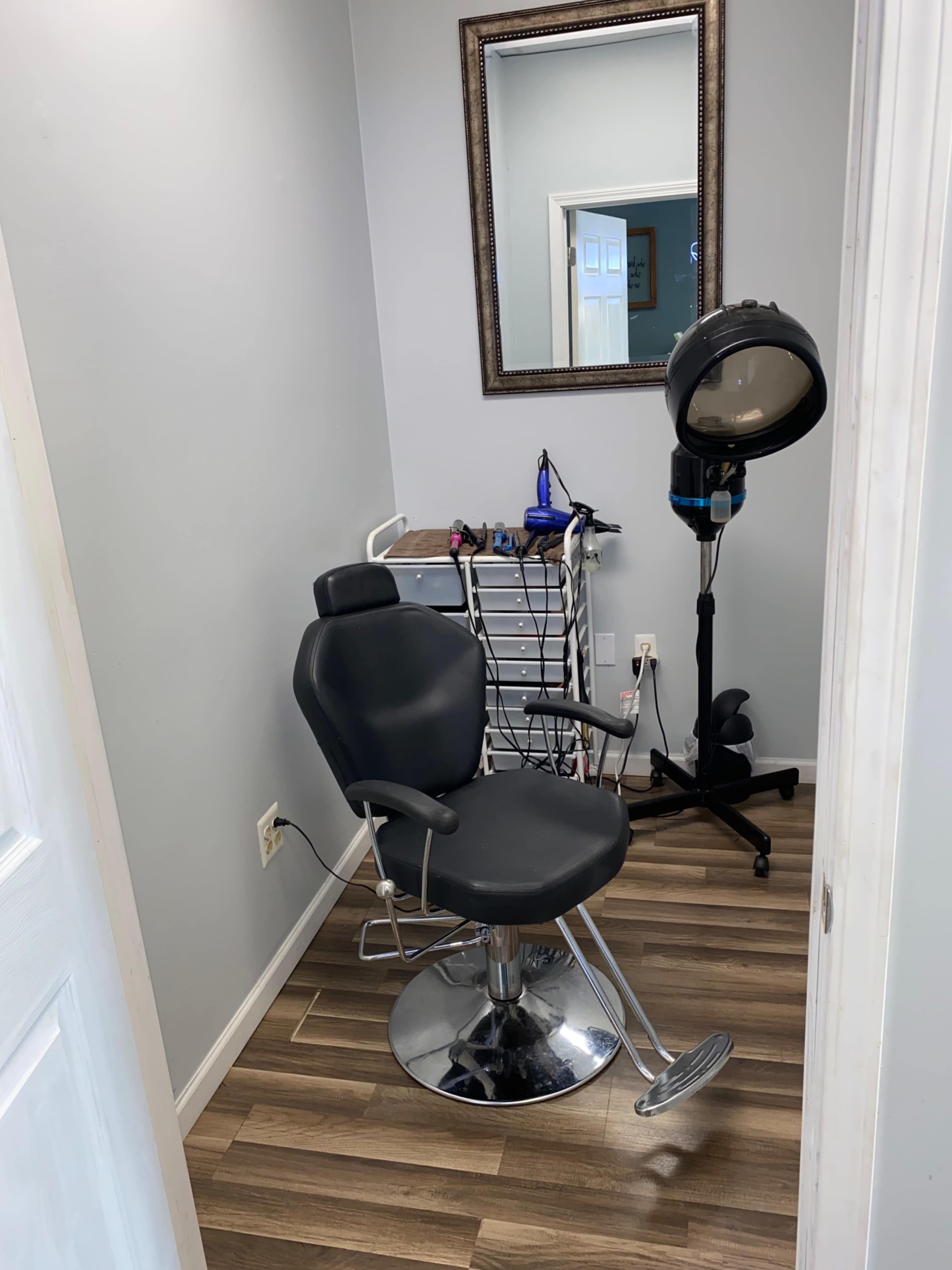 A hair salon corner featuring a black salon chair, a mirror on the wall, and a hooded hairdryer stand beside a rack with various hair styling tools.