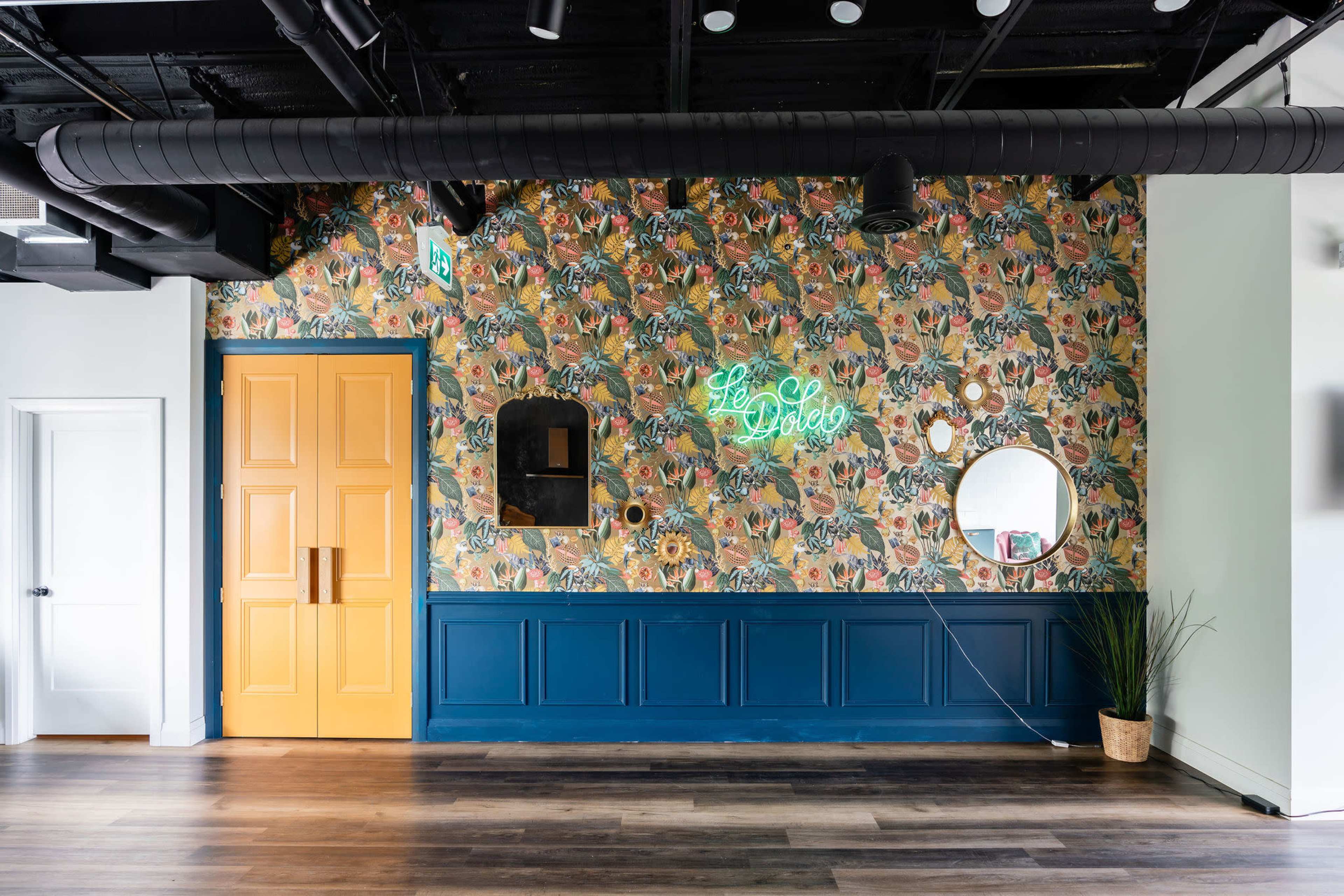 The image shows a brightly decorated wall with floral wallpaper, a yellow door, a circular mirror, and a neon sign, all set against a blue wainscoting.