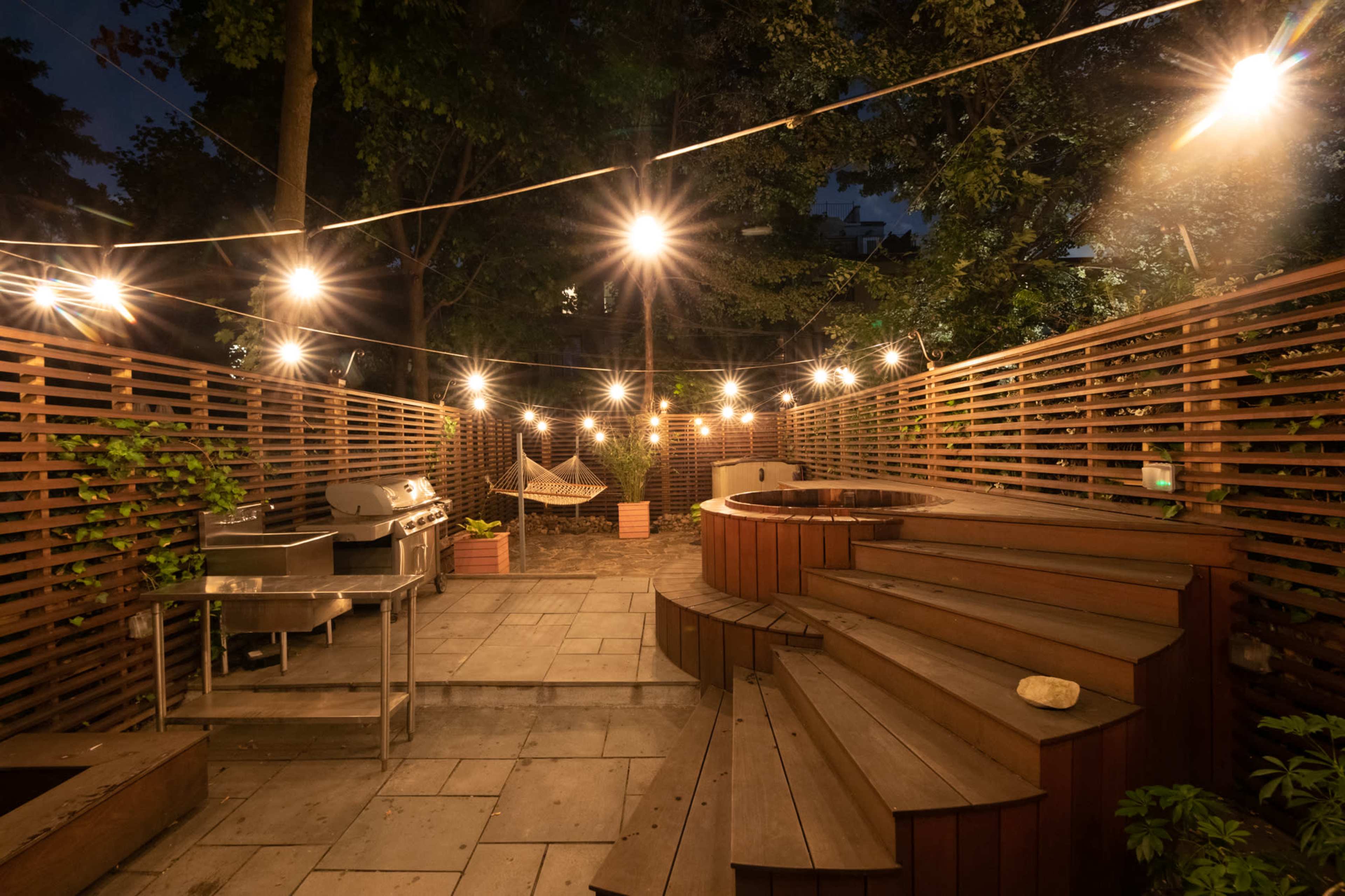 The image depicts a well-lit outdoor patio area at night, featuring a circular hot tub, wooden seating steps, a grill, and string lights overhead, surrounded by wooden fencing and potted plants.