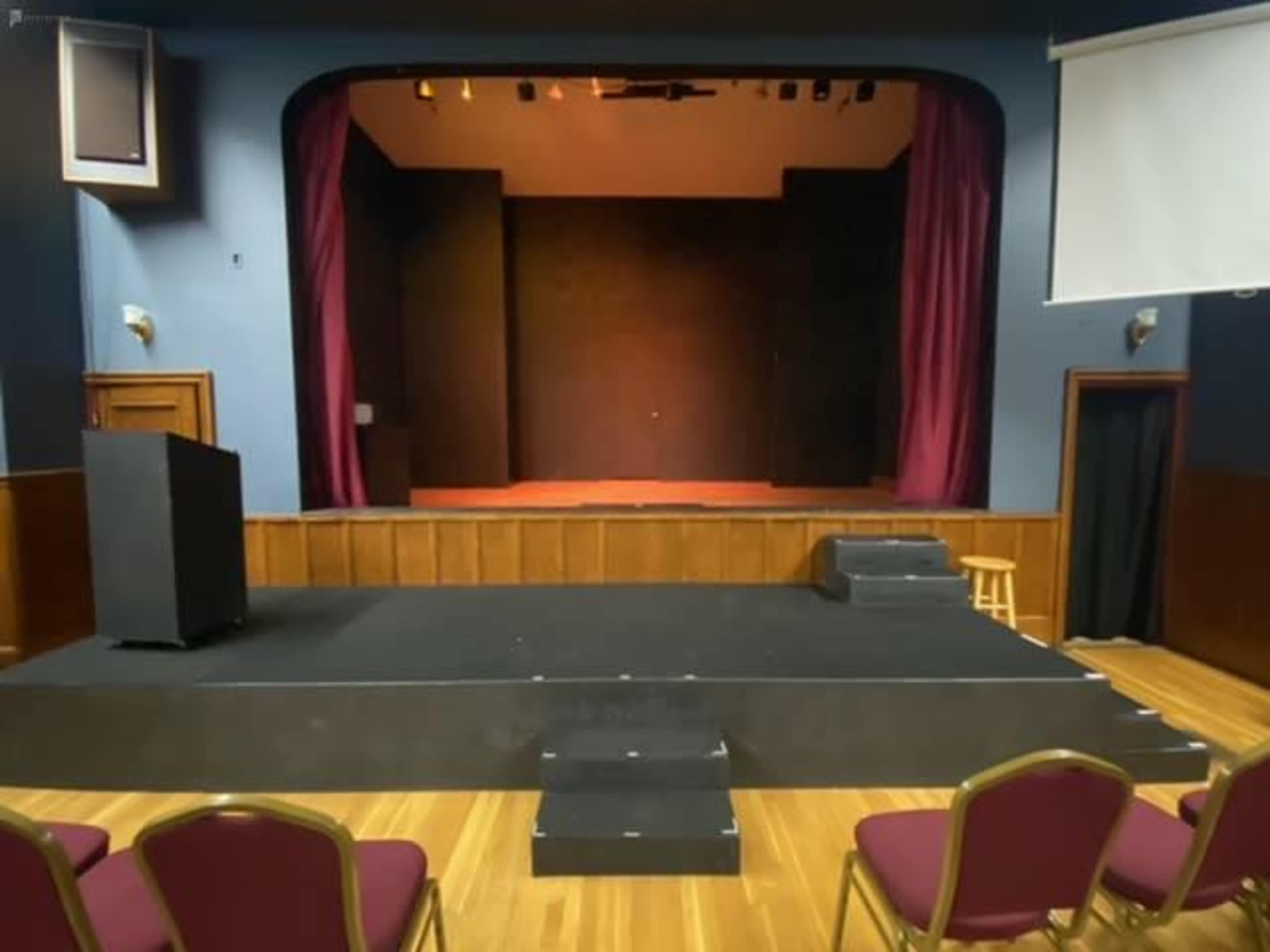 The image shows a small stage in a theatre, framed by maroon curtains, with a podium and several chairs arranged in front.