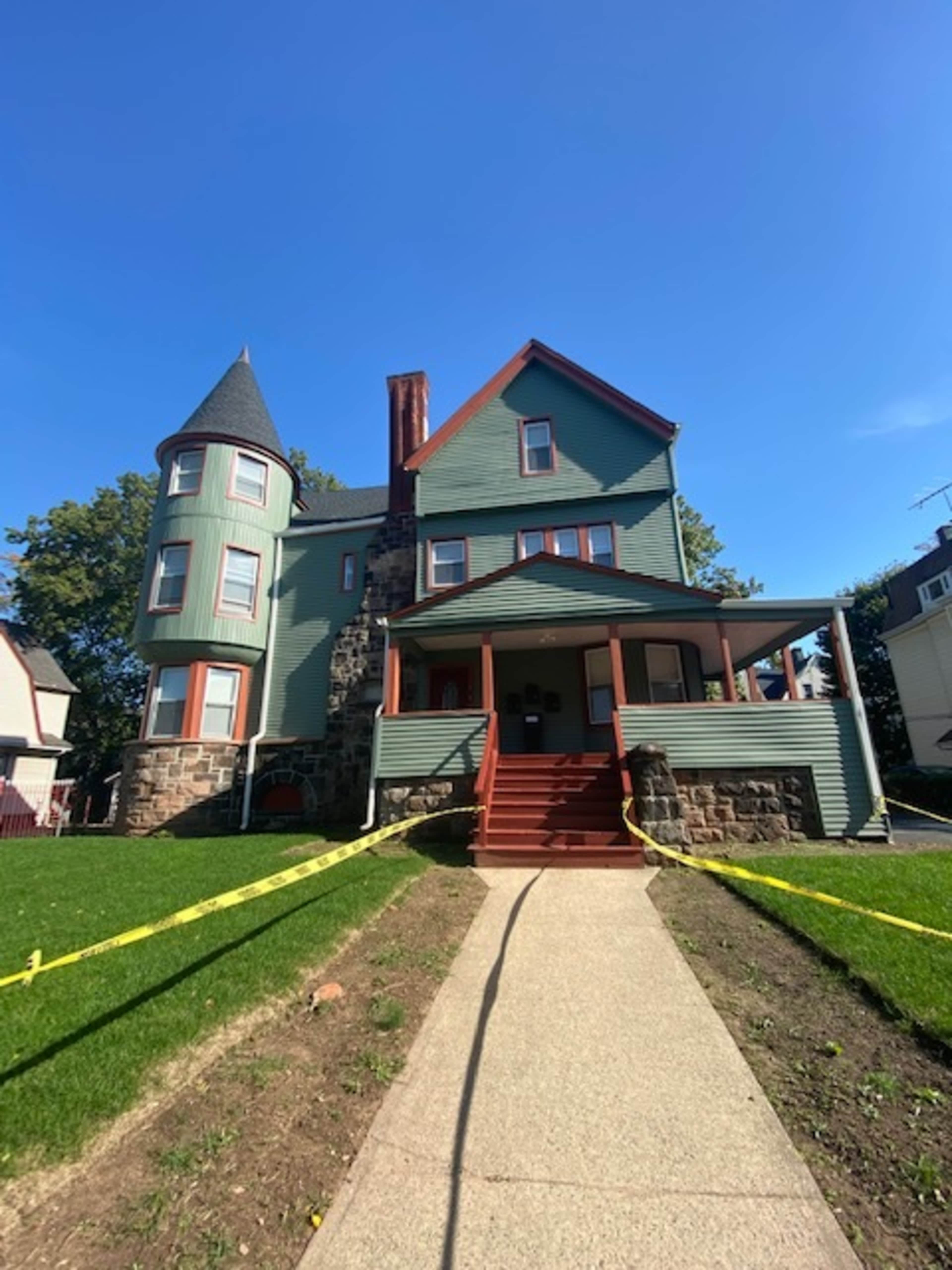 The image shows a multi-story house with green siding, a stone base, a turret, and a red staircase leading to the entrance, surrounded by yellow caution tape.