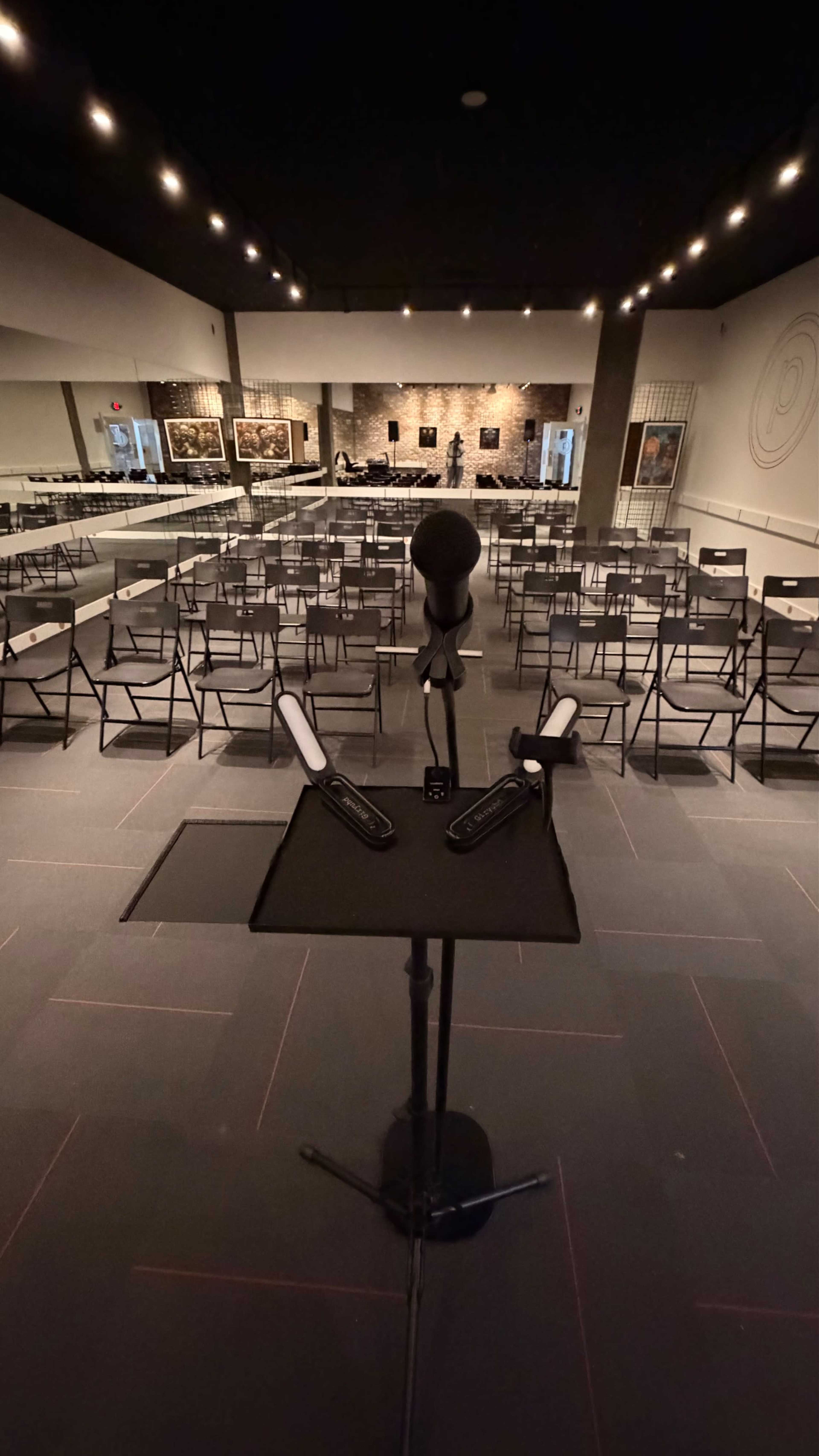 The image shows a microphone on a stand in the foreground with a seating area of folding chairs arranged in rows in a dimly lit room.