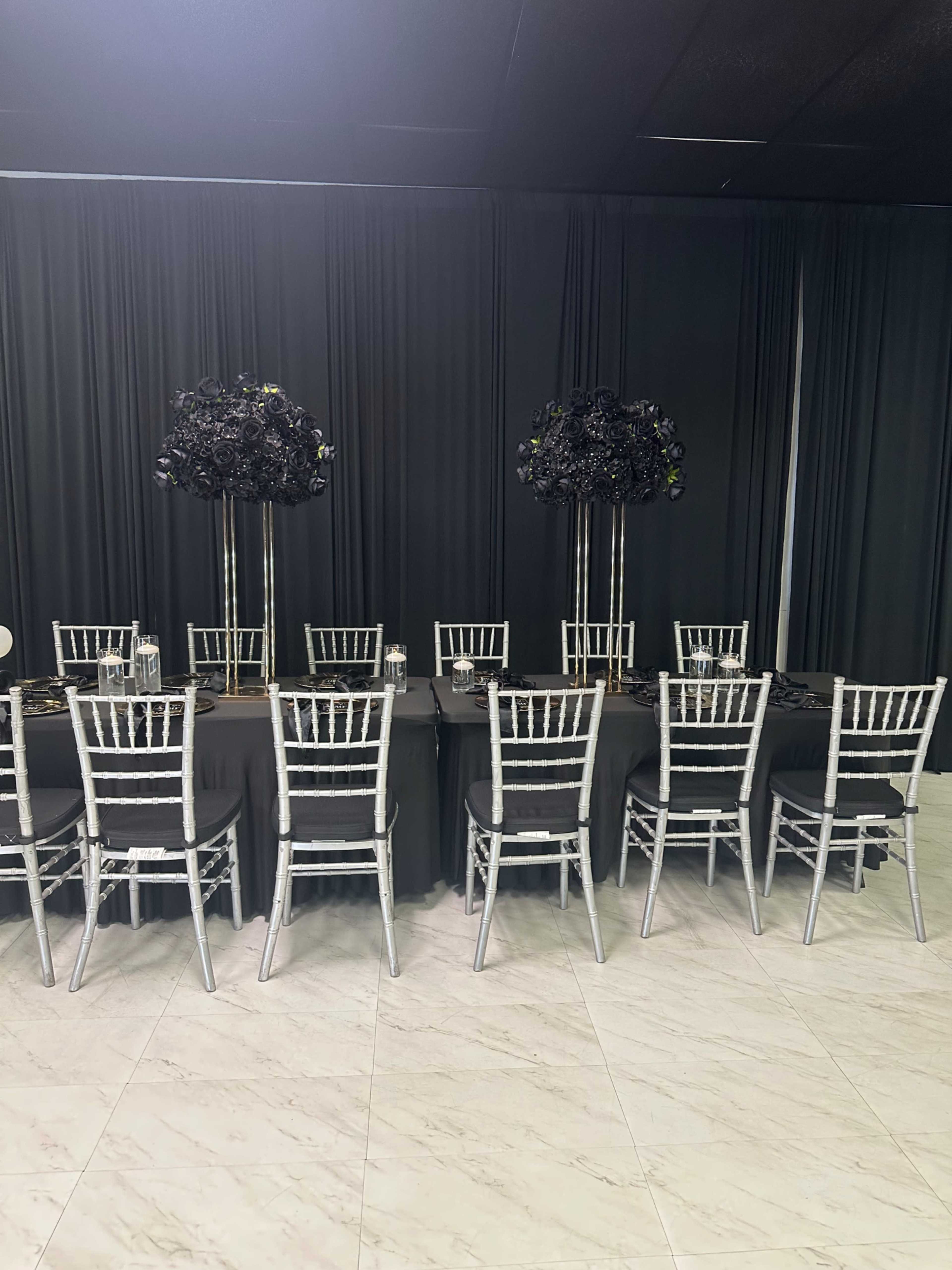 A long dining table is set with black tablecloths and silver chiavari chairs, featuring tall decorative arrangements on either end.