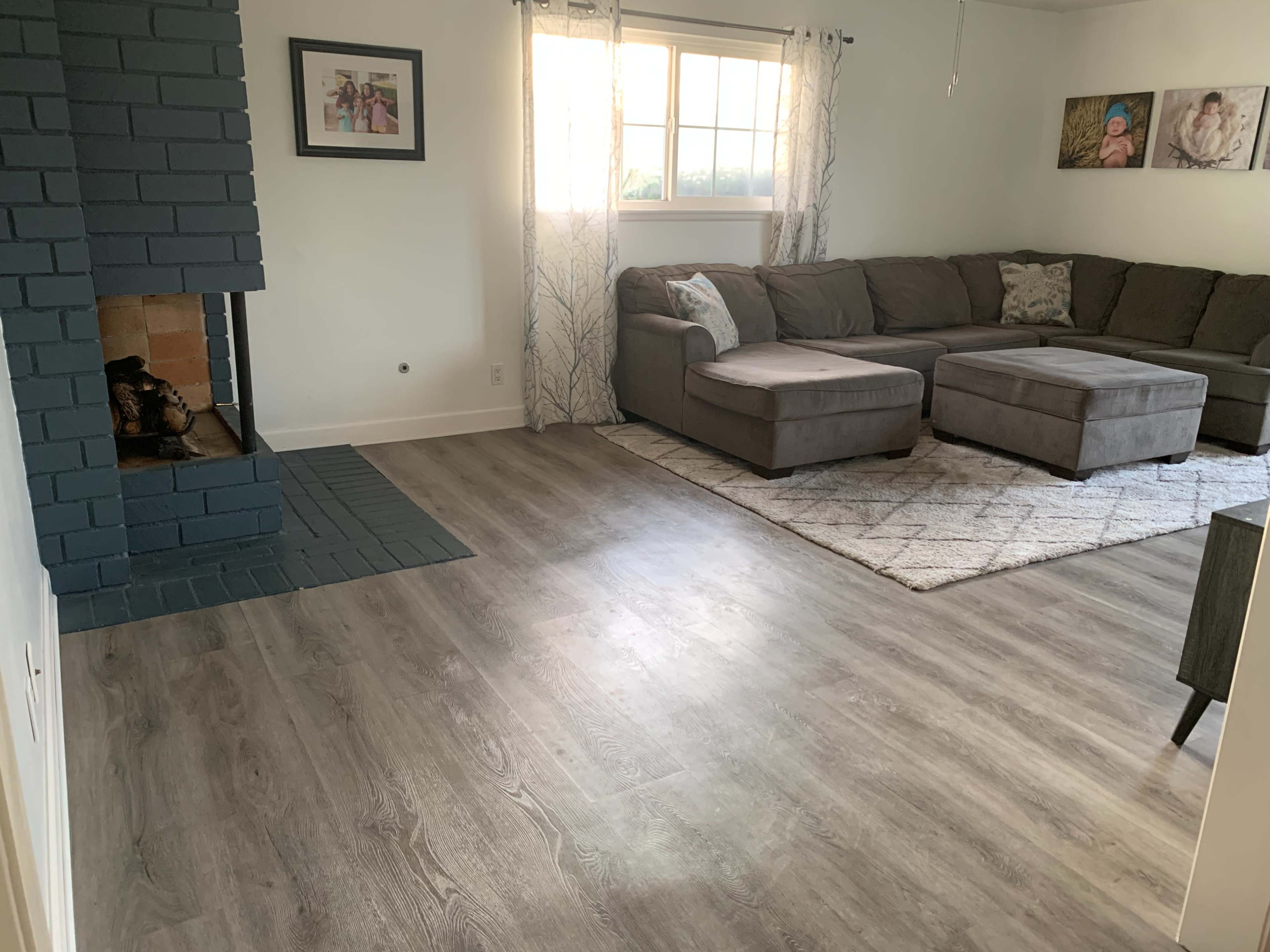 The image shows a living room with a gray sectional sofa, an ottoman, a fireplace with a blue-painted brick surround, and a window allowing natural light to enter the space.