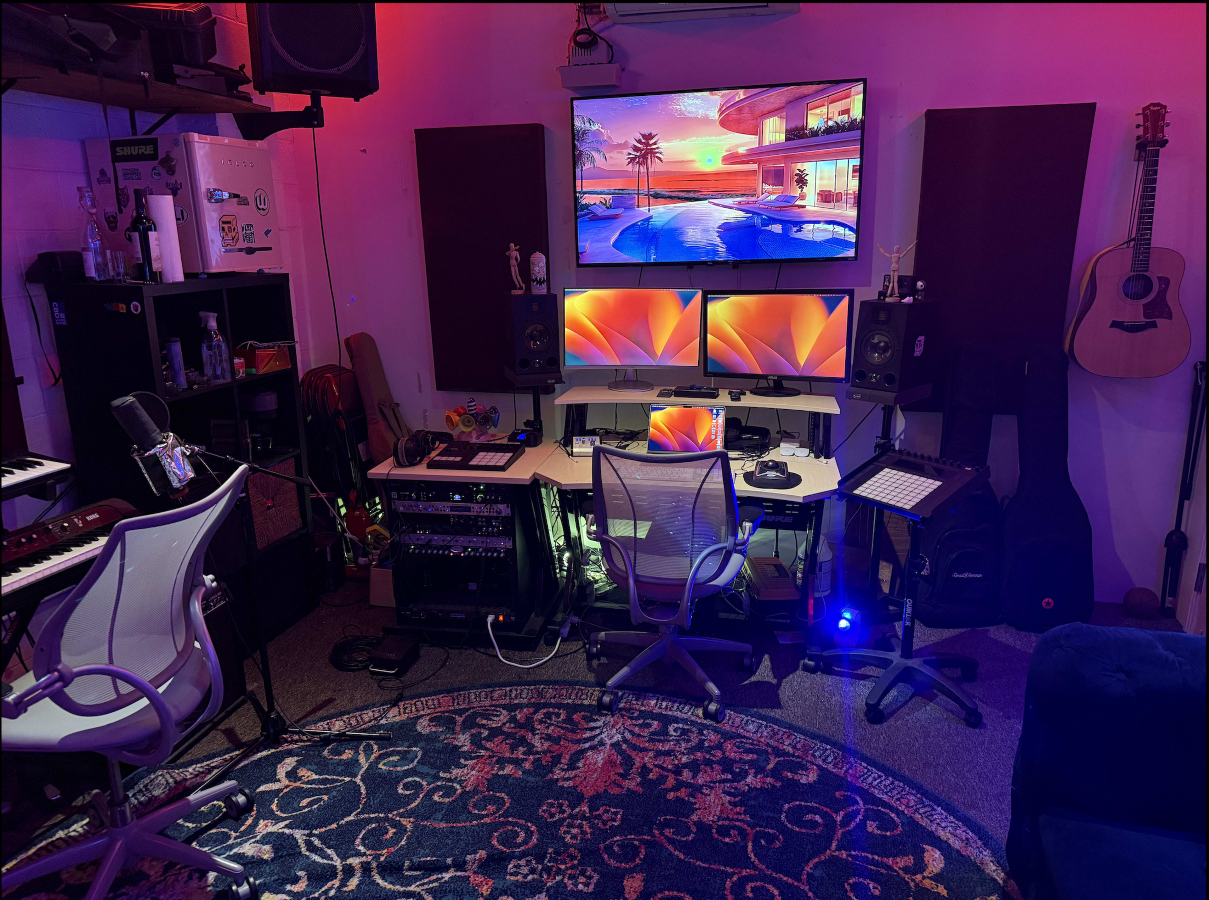 The image shows a music production studio with two monitors, audio speakers, a keyboard, and various instruments arranged in a compact space.