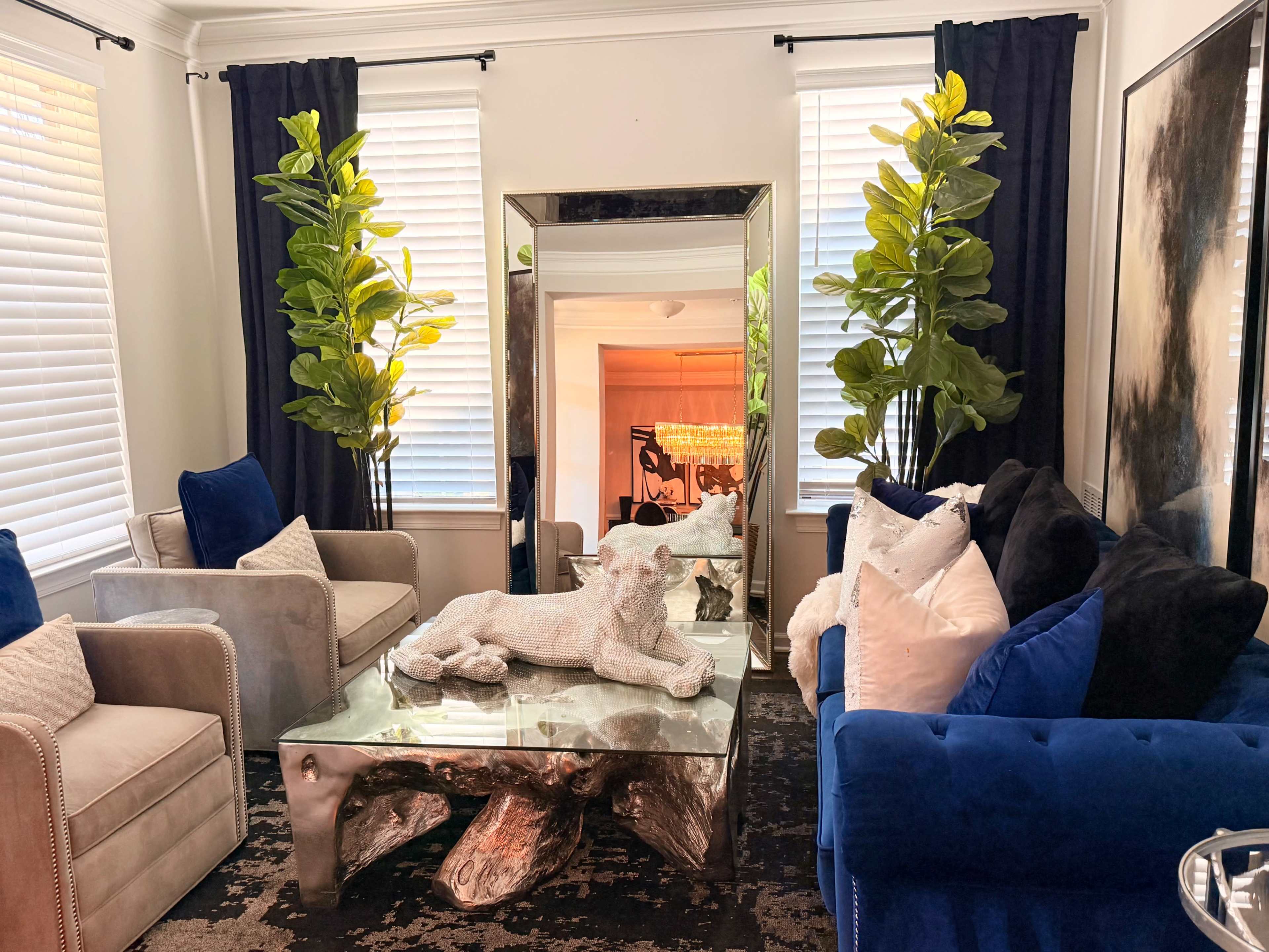 The image shows a stylish living room with two plants flanking a large mirror, a coffee table featuring a decorative object, and two sofas adorned with various pillows.