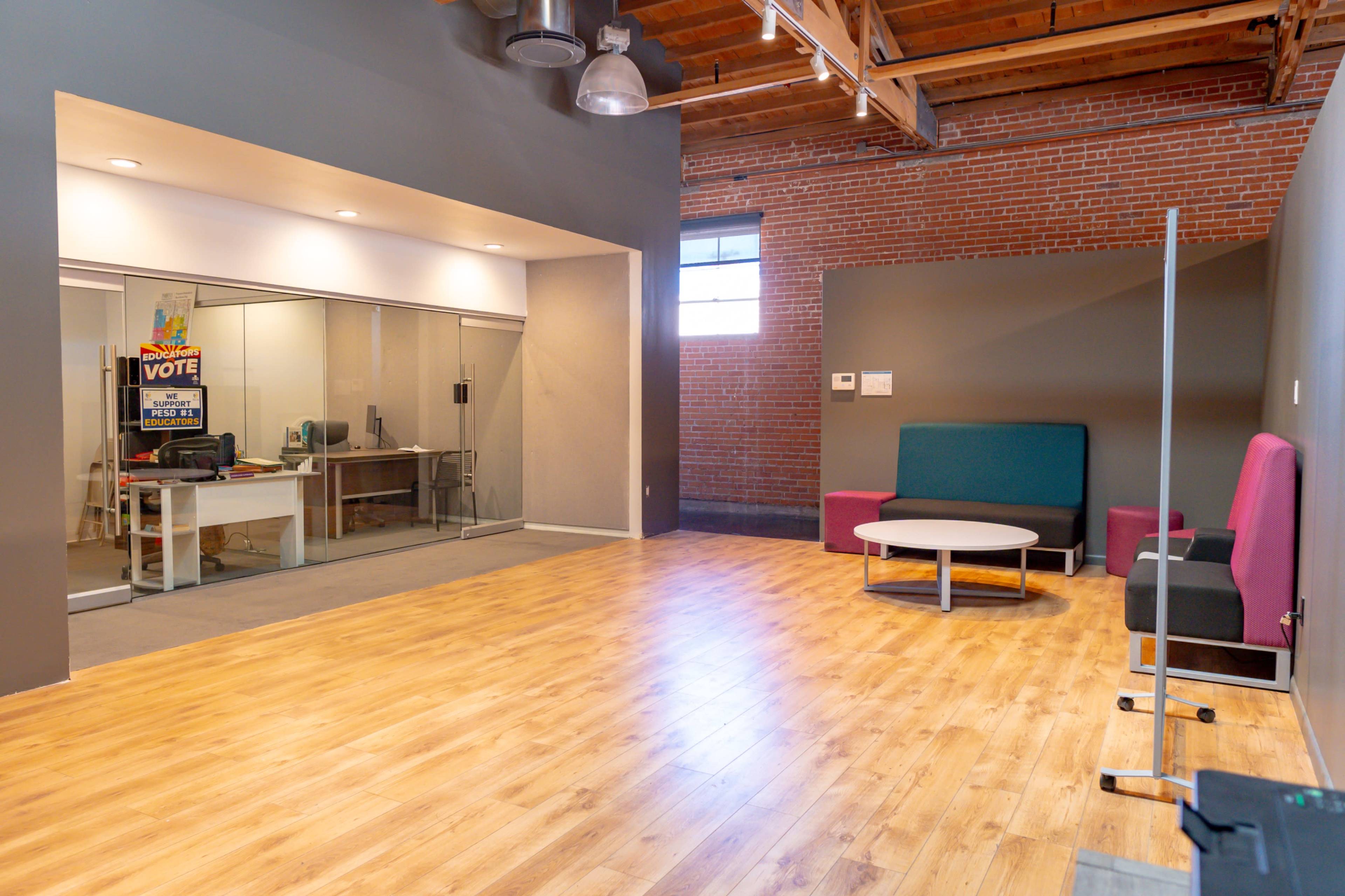 A spacious indoor area with wooden flooring, featuring a round table surrounded by seating, and a glass-walled office in the background.