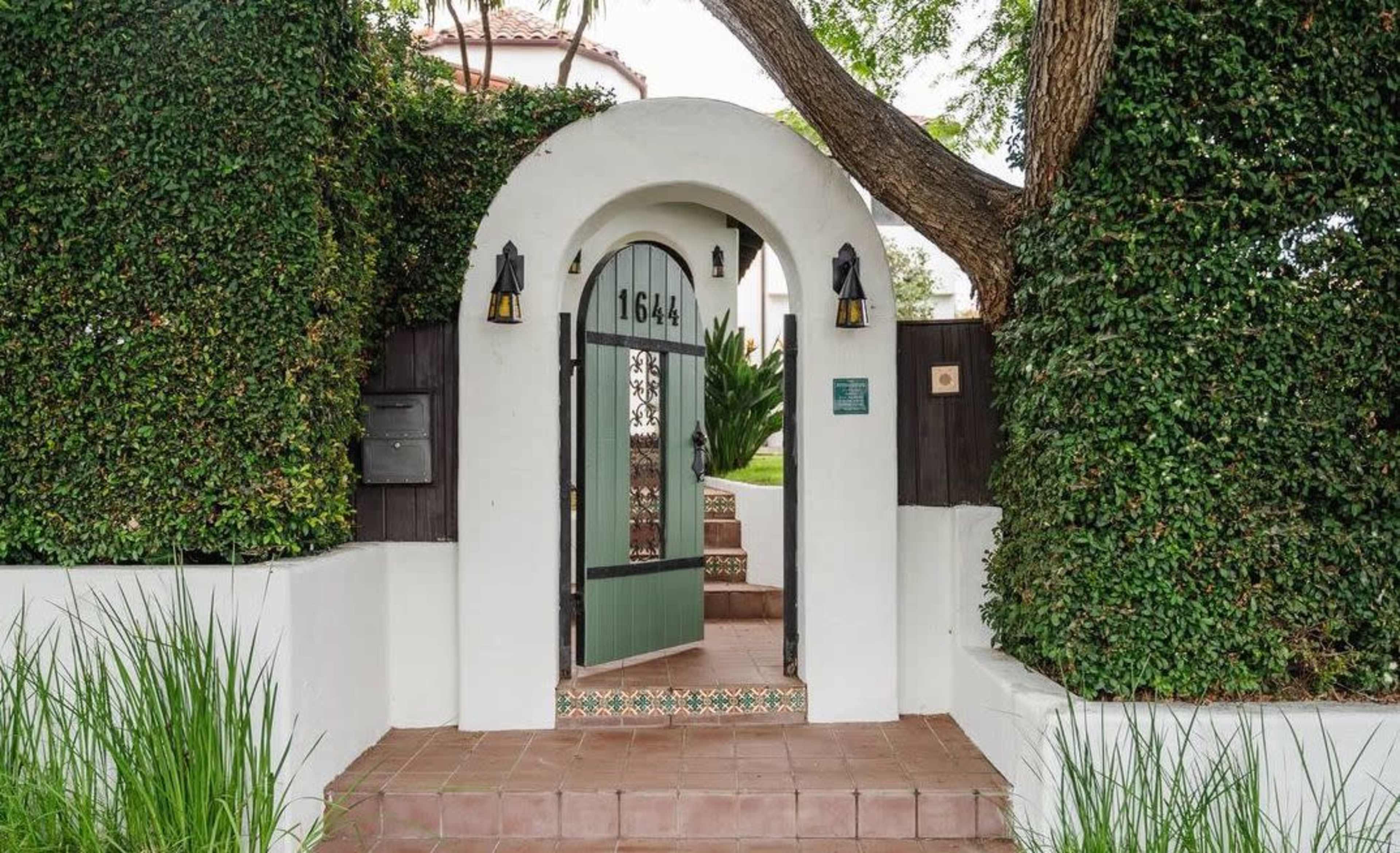 The entrance features a white stucco archway with a green door, surrounded by lush greenery and a tiled pathway leading to the home.