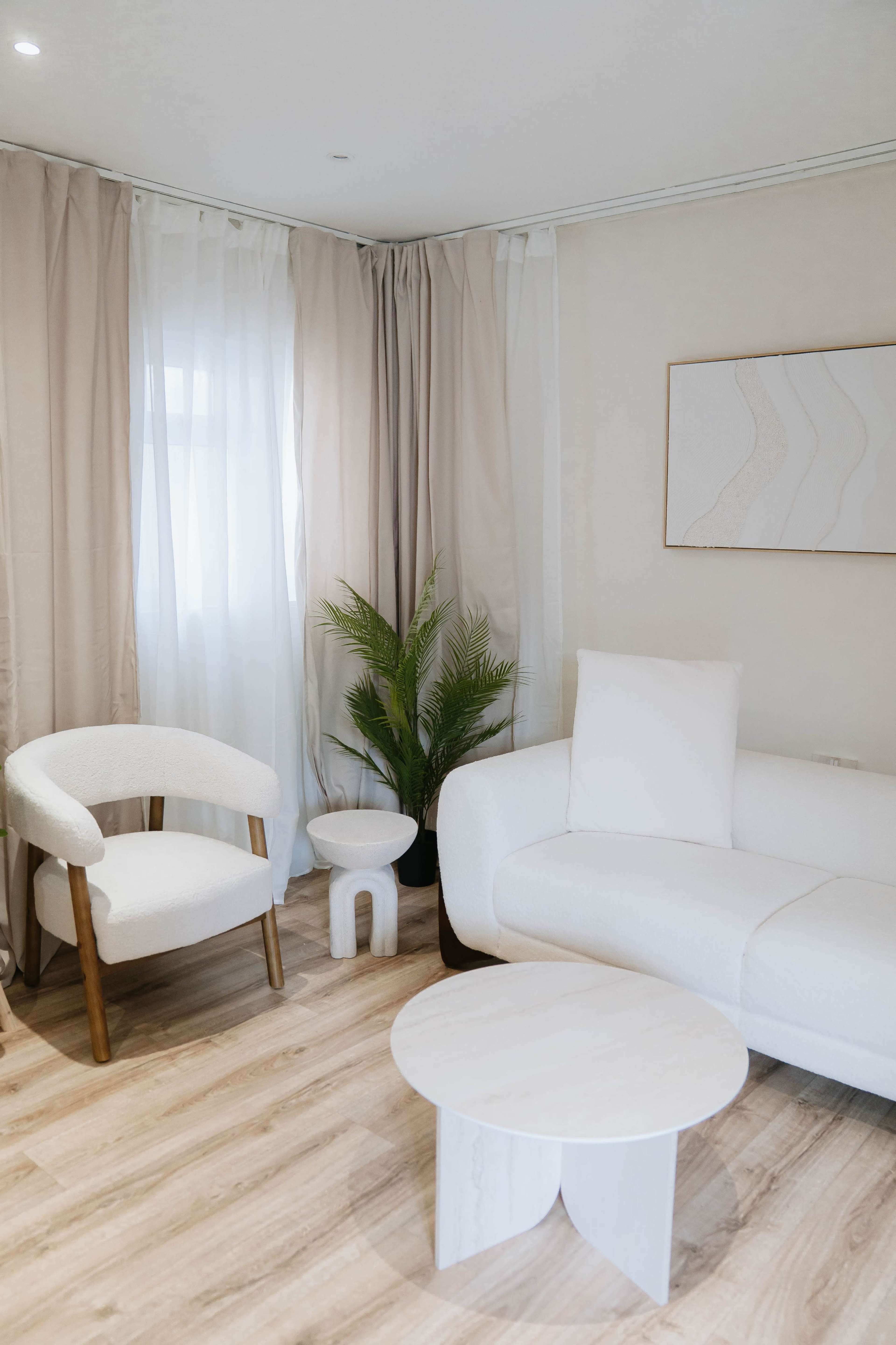 The image shows a modern, minimalistic living room featuring a white sofa, a round coffee table, a small side table, and a green plant, all set against light-colored curtains.