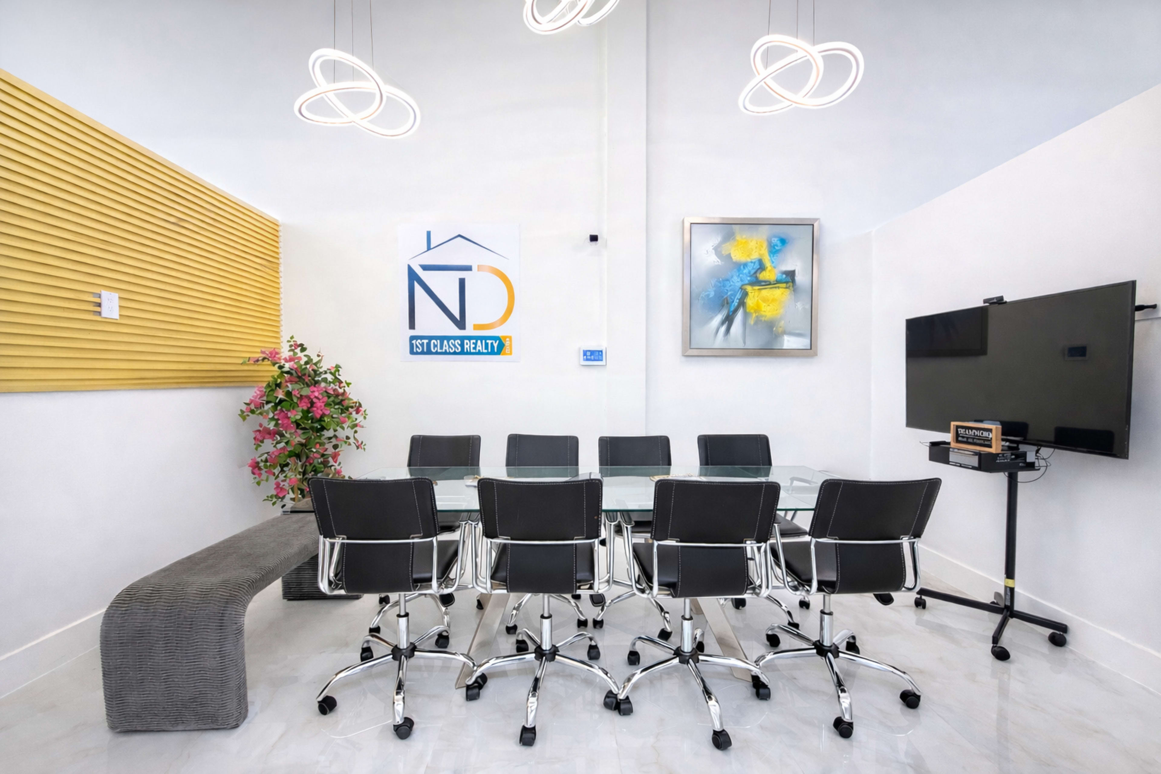 The image shows a modern conference room with a glass table surrounded by black office chairs, a large screen on a stand, and wall decorations including a logo and a painting.