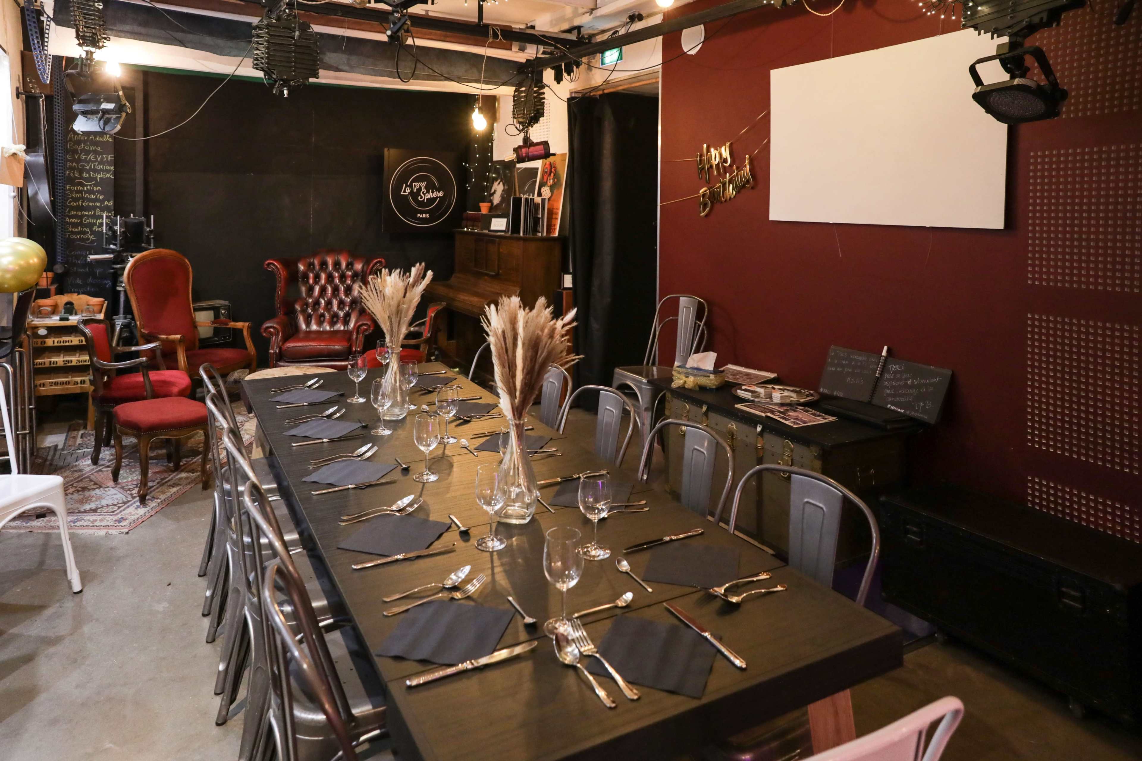 A dimly lit dining area with a long table set for a meal, featuring decorative vases and silverware, surrounded by eclectic furniture and a backdrop of red walls.