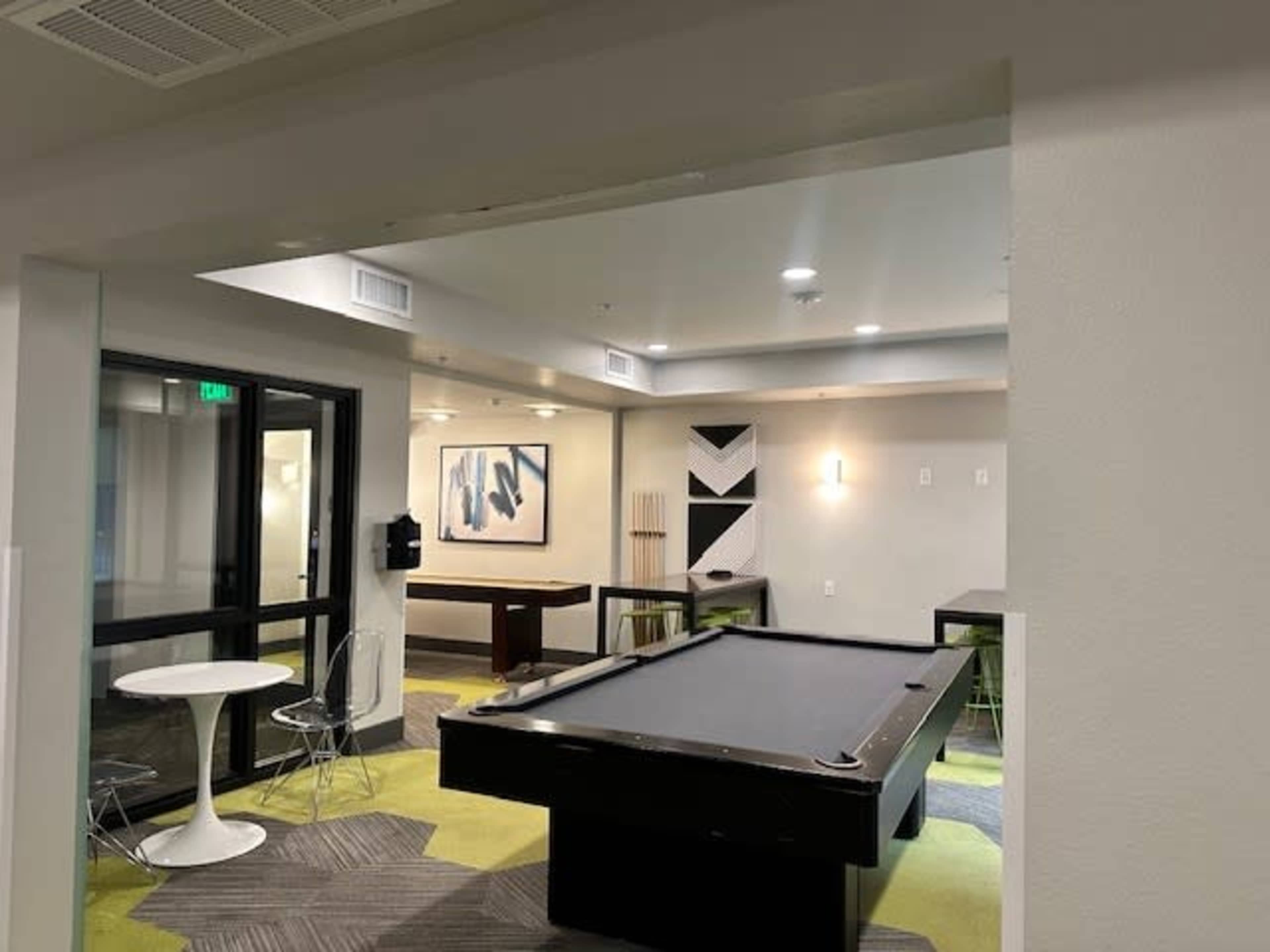 The image depicts a modern recreational room featuring a pool table, seating area, and art on the walls.