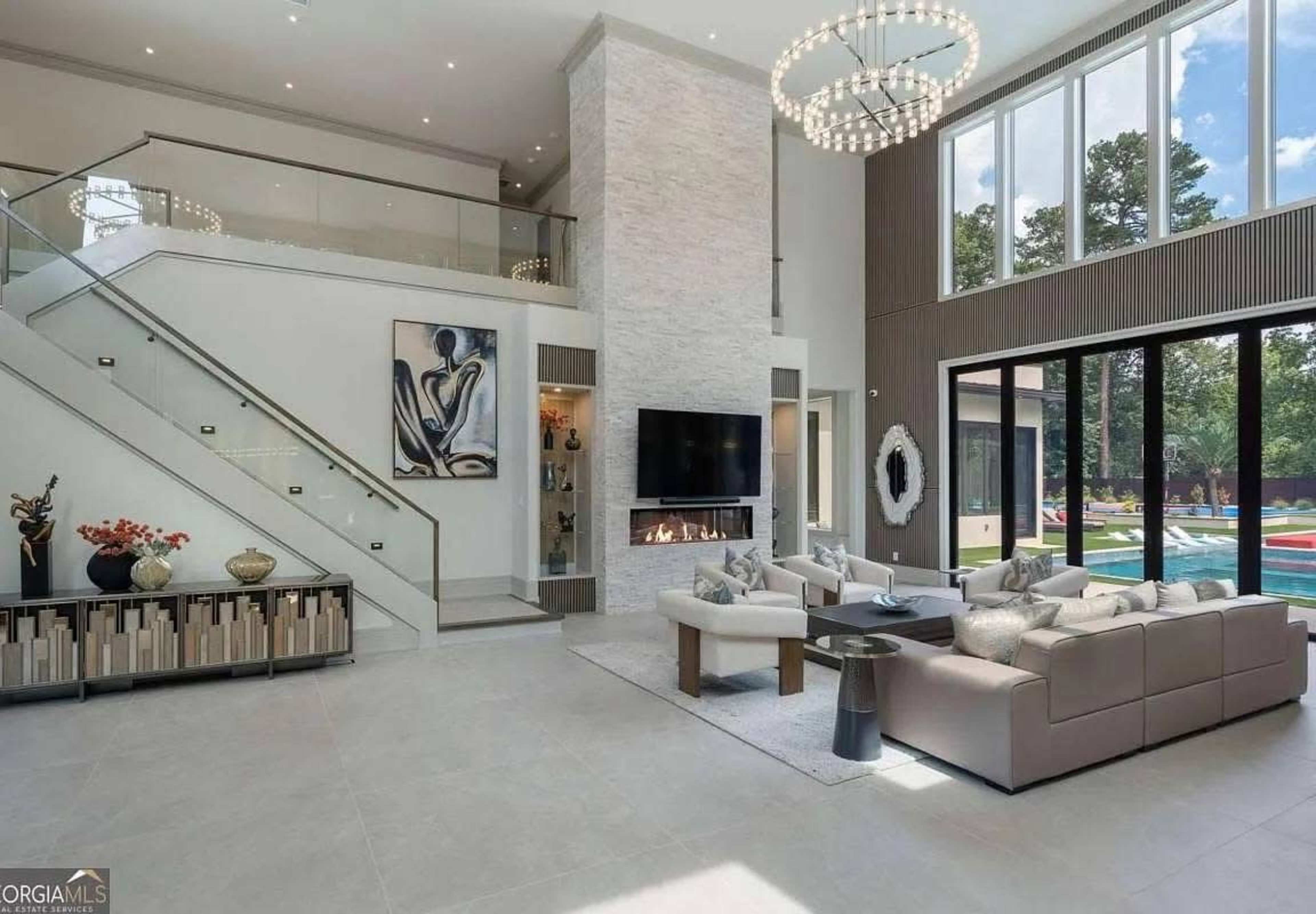 The image shows a modern living room featuring a sleek design, large windows, a fireplace, and a two-story ceiling.