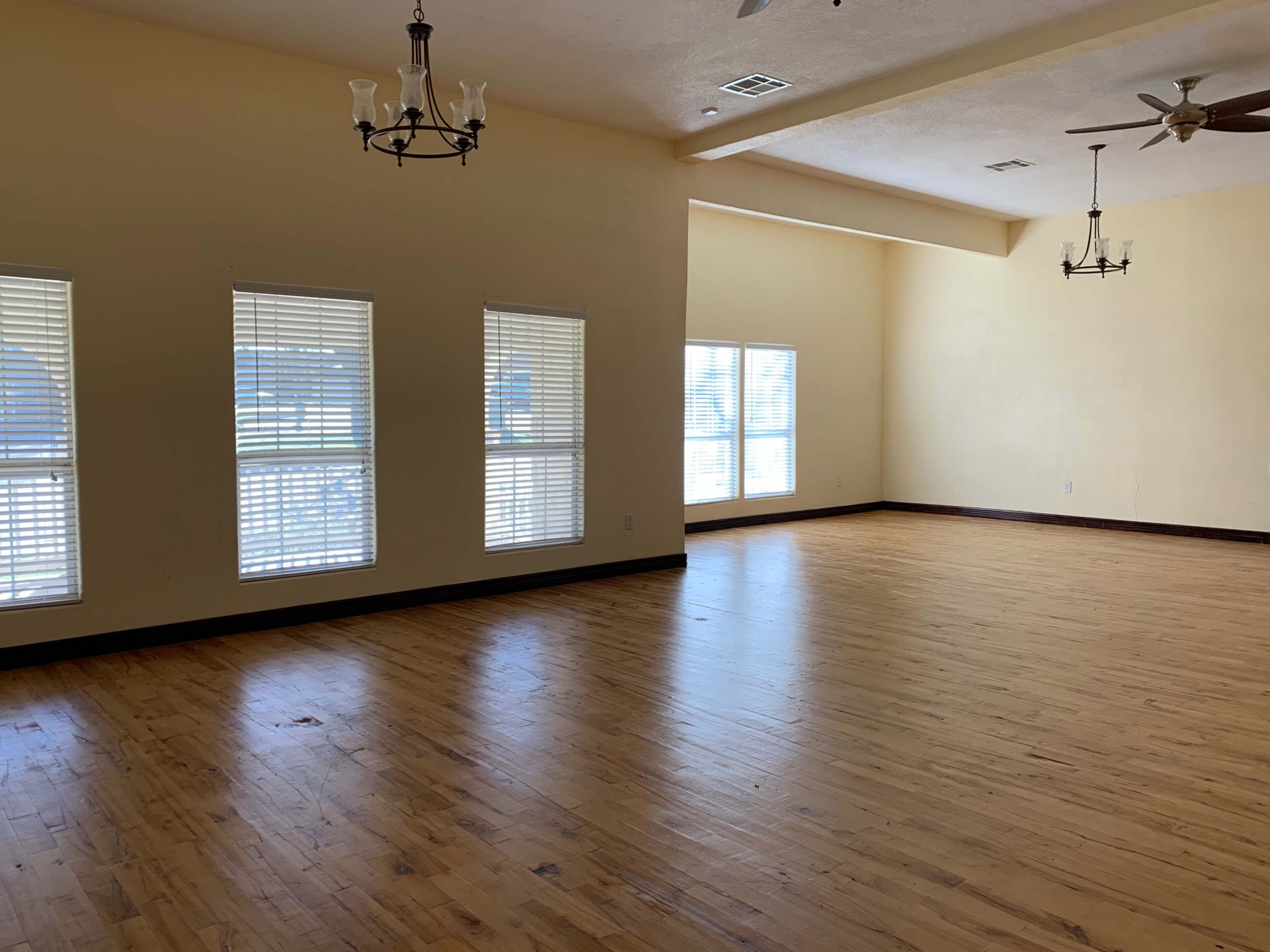 A spacious room with wooden flooring, large windows, and two ceiling fans.