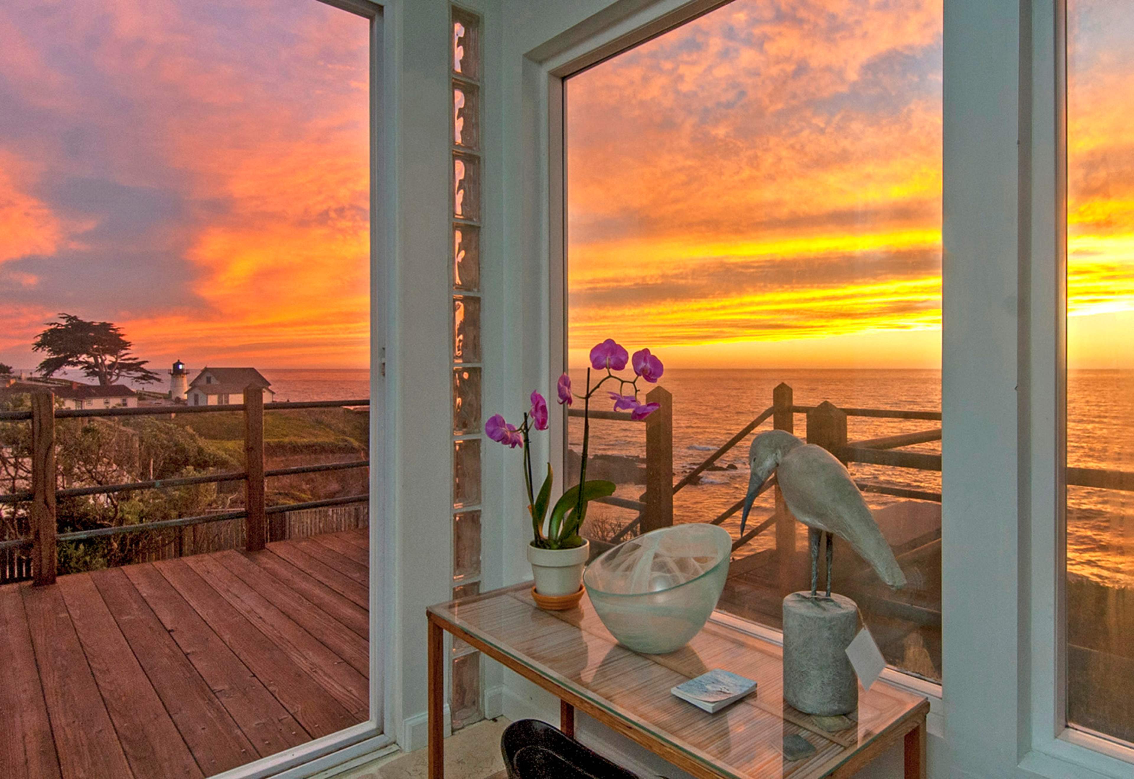 The scene features a coastal view at sunset through large windows, with a table displaying a vase of orchids and a decorative bird statue.