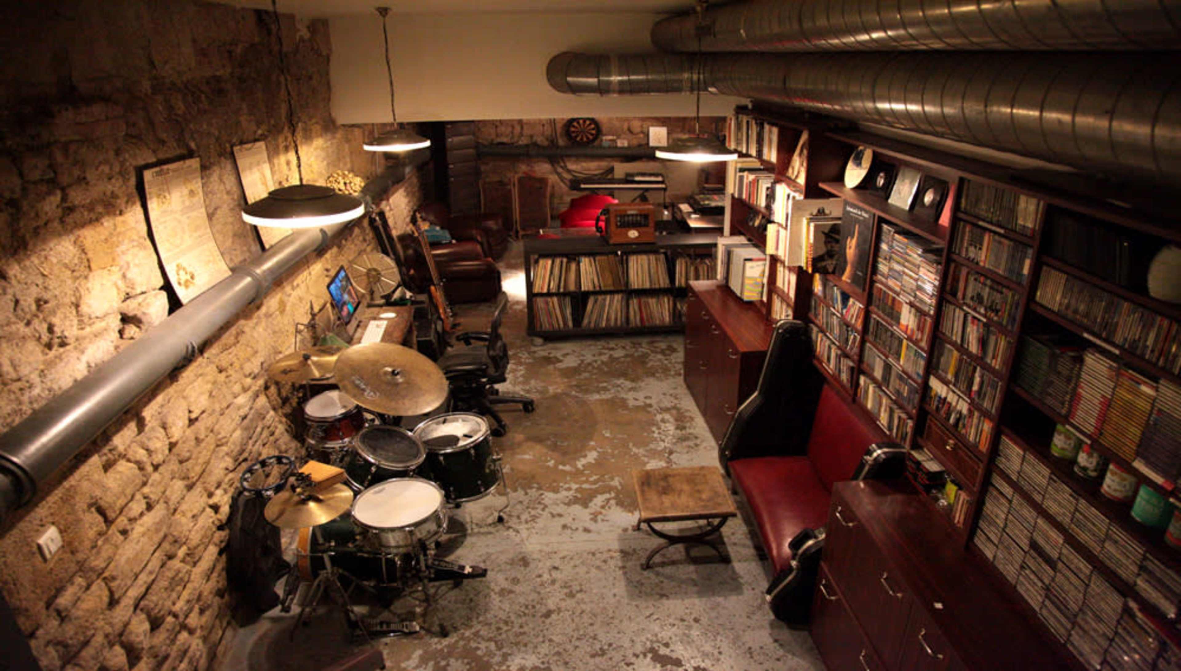 A music studio featuring a drum set, vinyl record collection, and various instruments arranged against a stone wall.