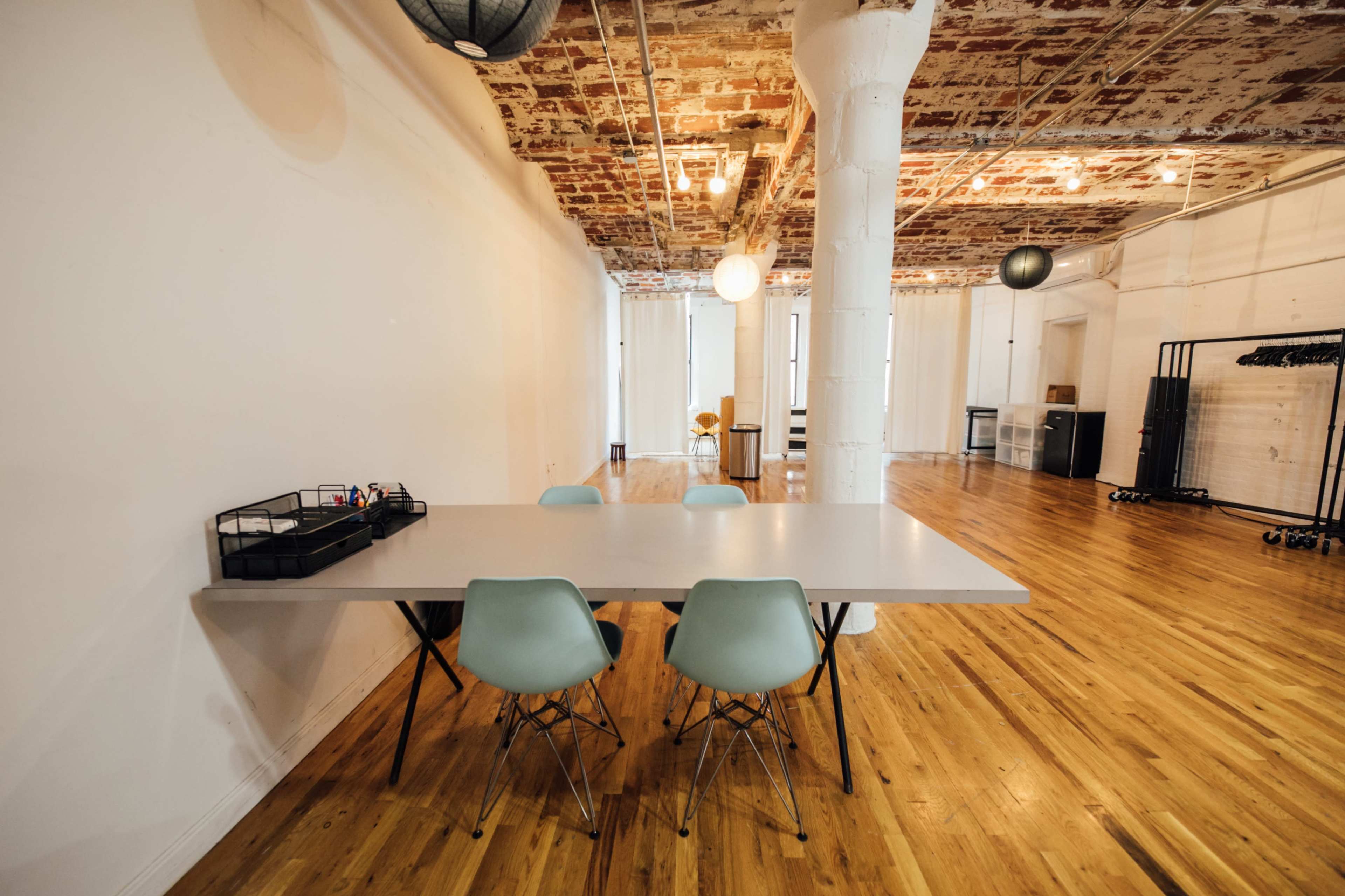 A spacious room features a large table with four light blue chairs, wooden flooring, and exposed brick walls.