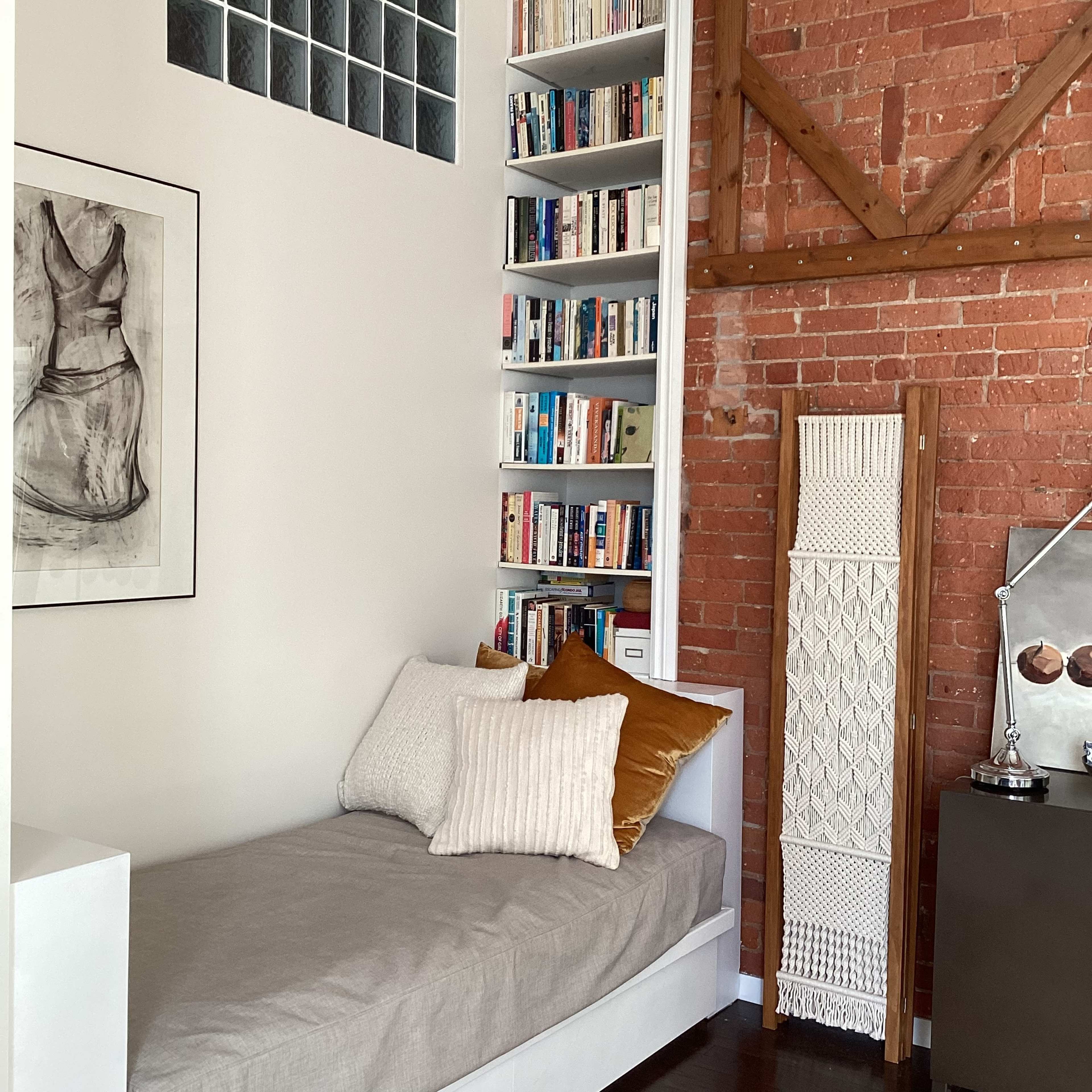 A cozy corner of a room featuring a bed with pillows, a tall bookshelf filled with books, and exposed brick walls.
