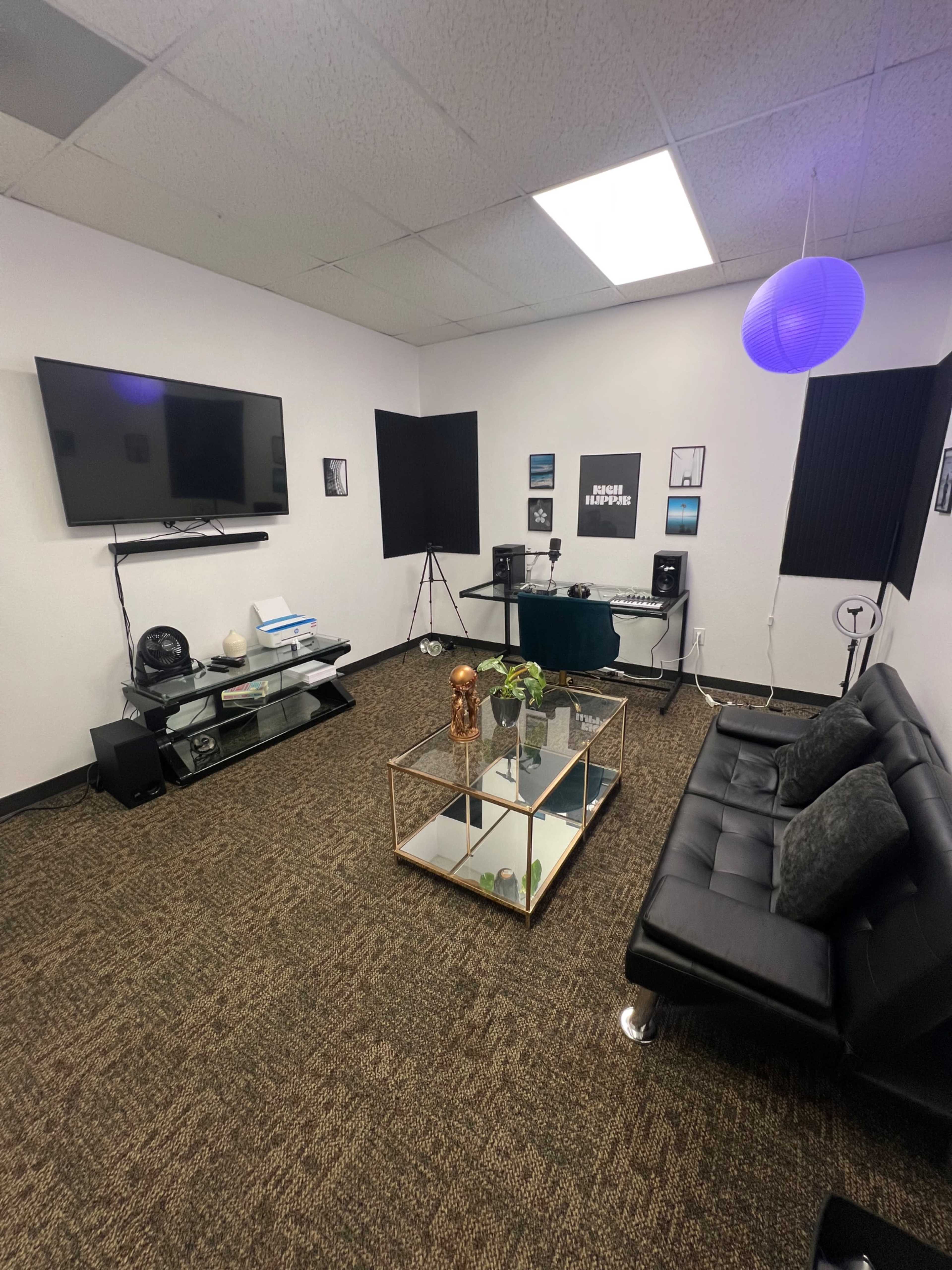The image shows a modern, minimalistic studio room with a black sofa, a glass coffee table, and audio equipment, along with a large television mounted on the wall.