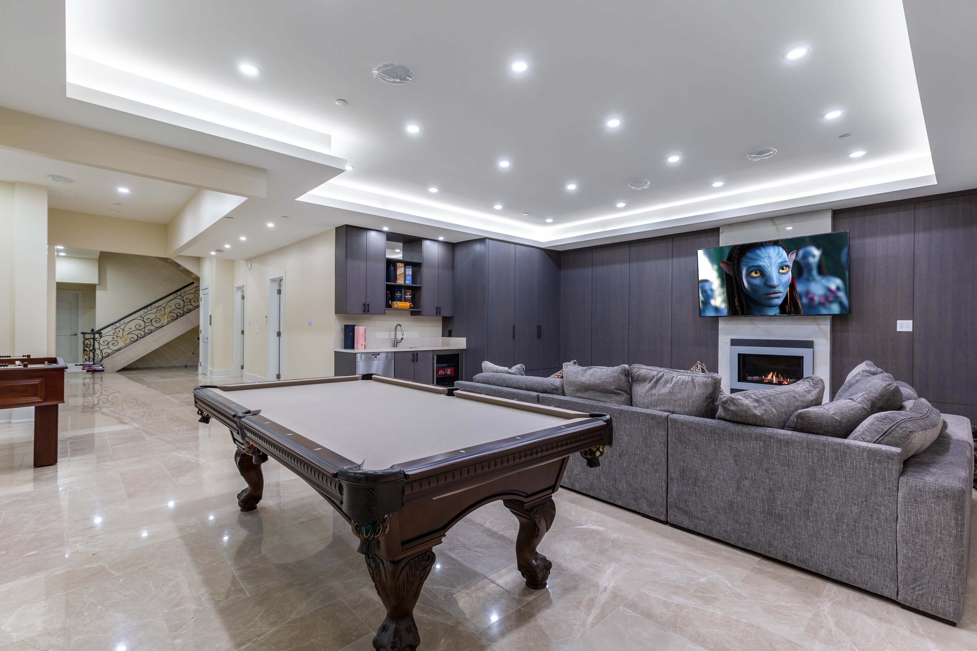 The image shows a modern entertainment room featuring a pool table, a large sectional couch, a wall-mounted television displaying an animated scene, and a sleek kitchenette area.