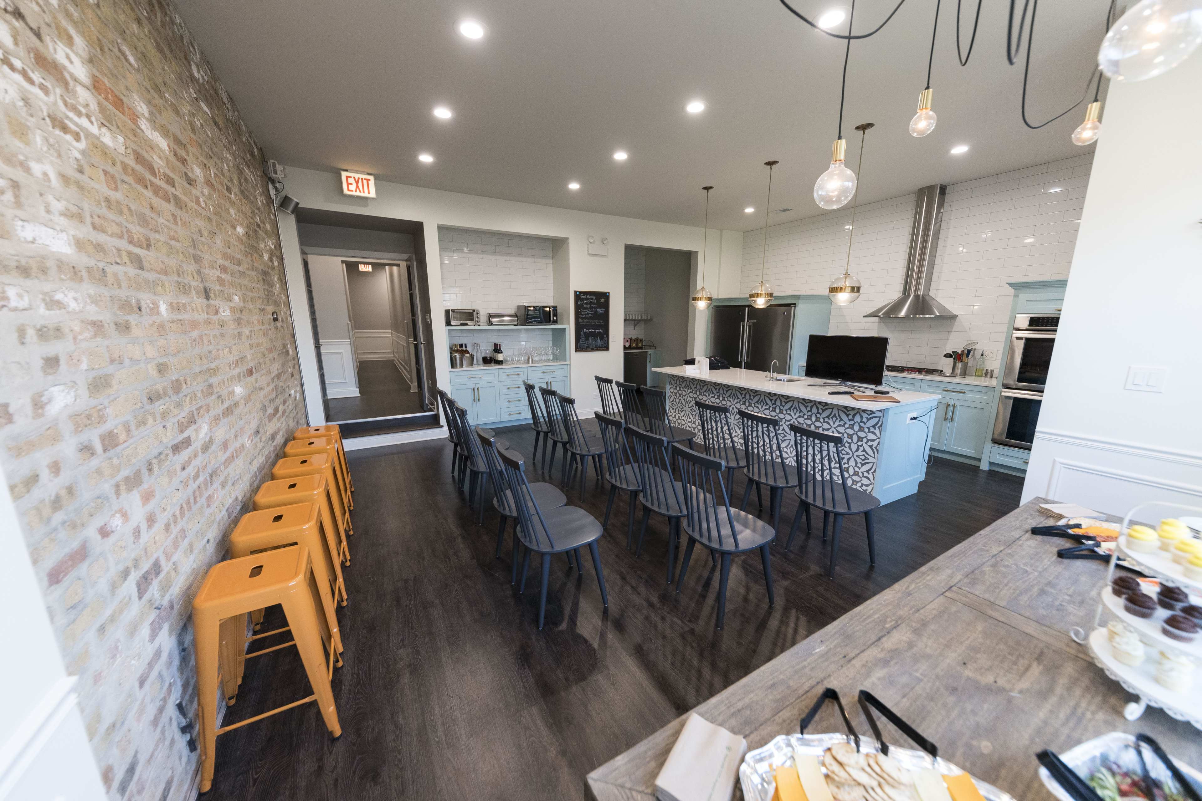 The image shows a modern kitchen and dining area with a seating arrangement of black chairs and orange barstools, featuring a brick wall and a well-equipped kitchen setup.